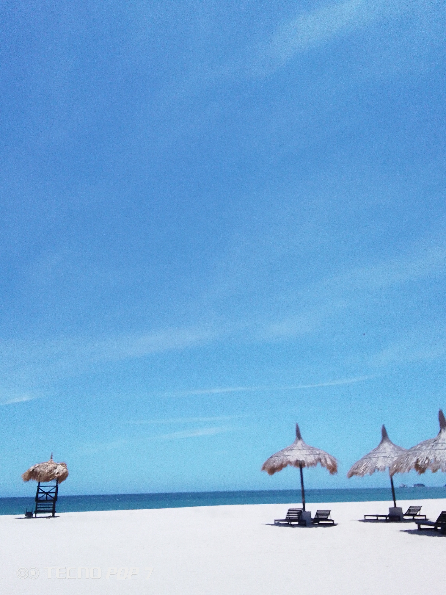 The image depicts a serene beach scene under a clear blue sky. The beach appears to be in Pedro González, Venezuela, evidenced by the local architectural design of the straw sunshades. The foreground is composed of white sand, gently sloping towards the calm blue sea.
There are four thatched-roof sunshades. One, situated on the far left of the frame, appears to be atop a lifeguard chair. The other three are each accompanied by two beach chairs. There are no people visible in the frame. The sky occupies a large portion of the image, showcasing a bright blue hue with faint wisps of clouds.