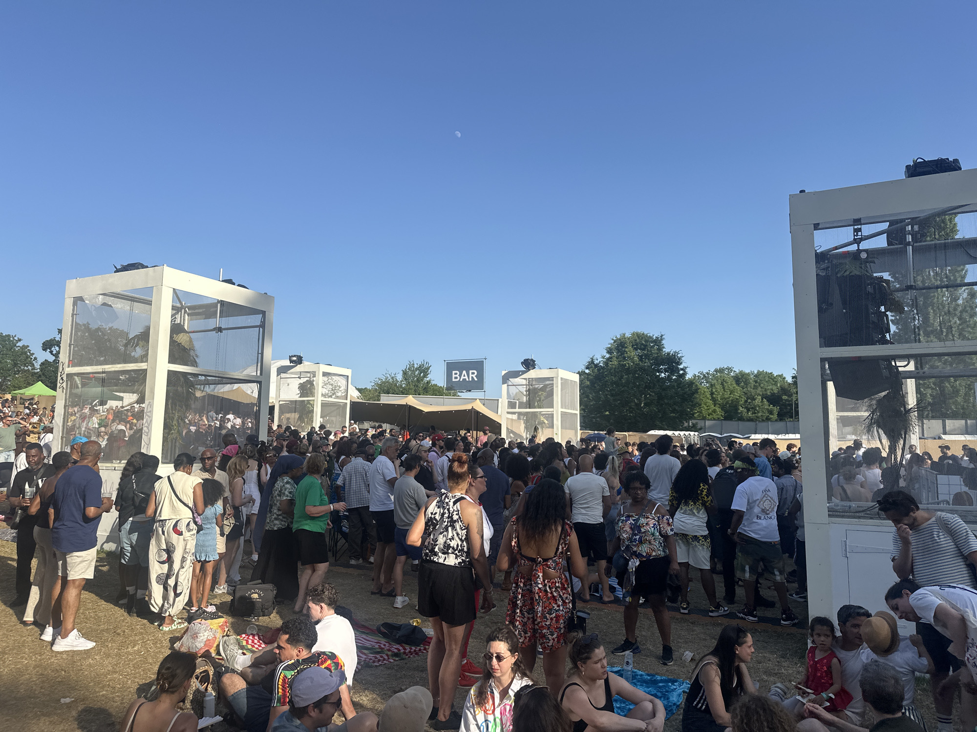 A large crowd of people is gathered in a park under a bright blue sky with a visible moon. Many people are standing and seem to be queuing, while others are sitting on the ground. Structures with white frames and transparent walls are present, along with a sign that says "BAR". The location appears to be an outdoor event in a green space, potentially in the city of London, United Kingdom.