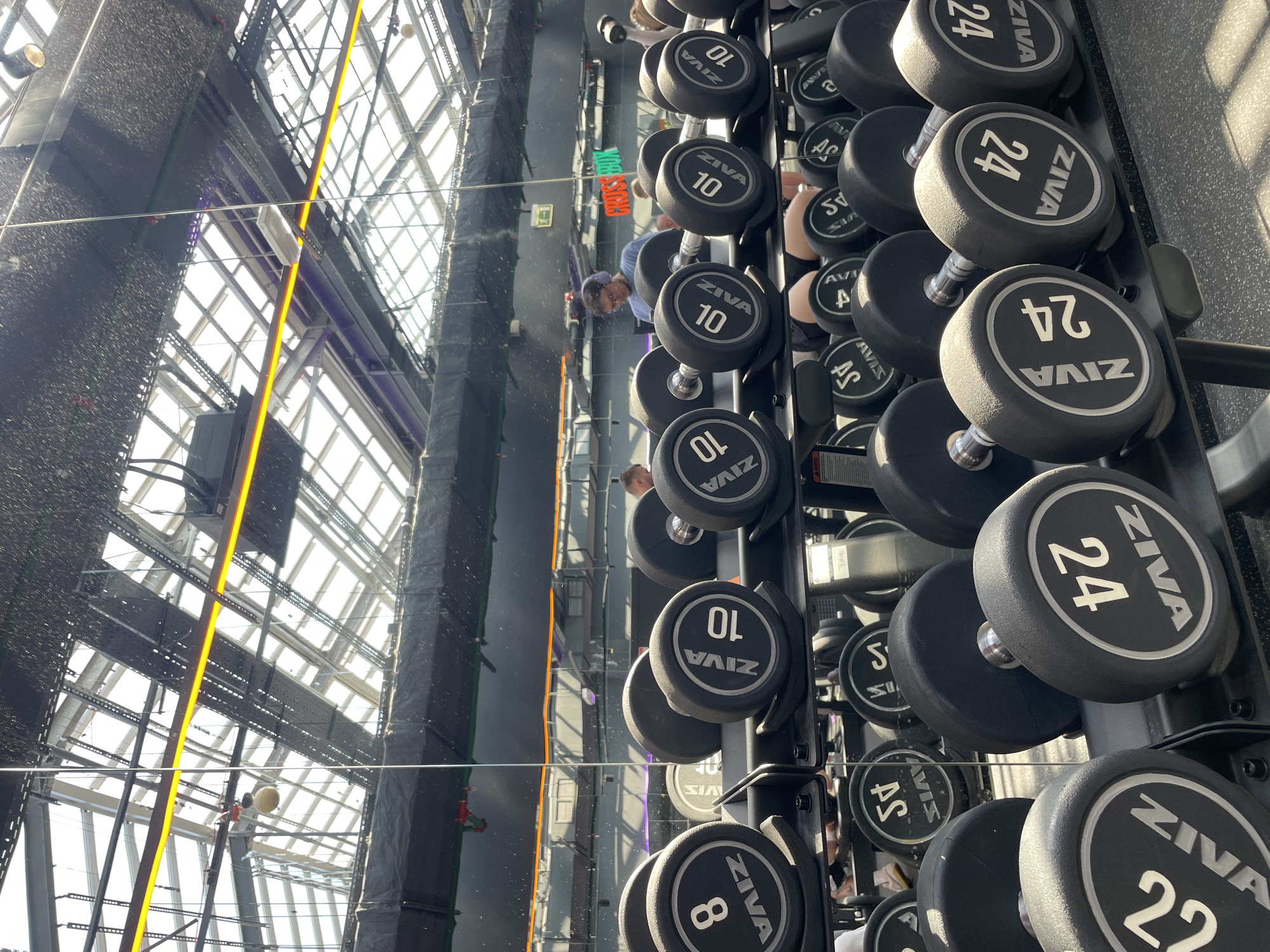 The image shows a dumbbell rack in a gym. The dumbbells are arranged in a neat and organized manner, ranging from 8kg to 45kg. The brand ZIVA is prominently displayed on each dumbbell.
The background reveals a modern architectural design, featuring a glass facade and a complex network of steel beams and supports. This architectural style is indicative of a modern sports facility or a gym located within a contemporary building.
Based on visual cues and the gym's architecture, there is a high probability that this photo was taken at a Cross Box gym in Kazan, Russia. This location is based on the "CROSS BOX" text visible in the reflection of the mirror, and the architectural style of the building, which is typical of modern sports facilities in Kazan.
The mirror also shows a reflection of individuals, giving the impression of people in the gym.