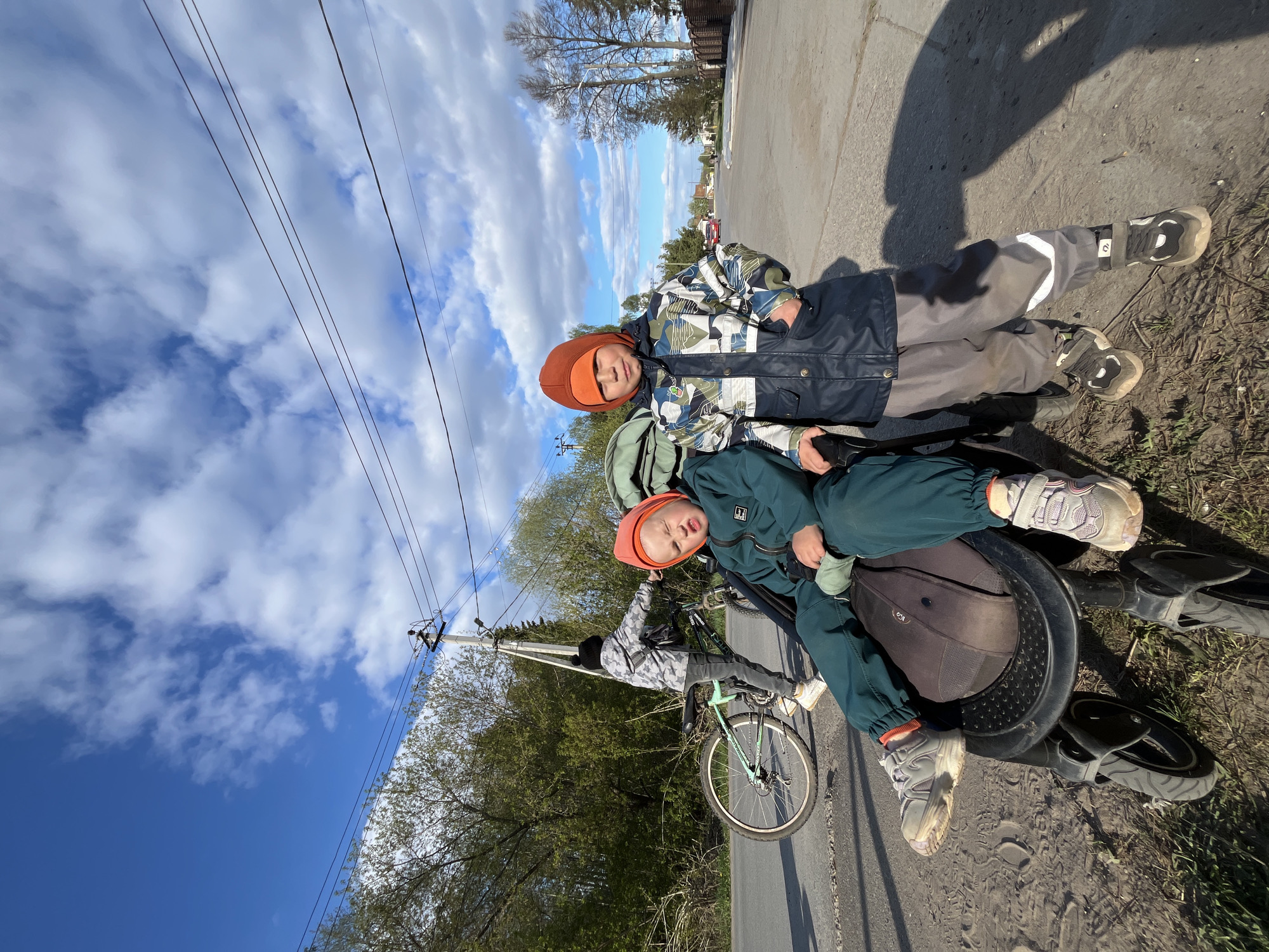 Two young children wearing winter hats and clothing are lying on a bicycle parked on the side of a road. The road is paved and abuts a patch of grass and trees. A utility pole with power lines is visible in the background. The sky is partly cloudy with blue patches. The photograph was taken in the city of Chaikovsky, Russia.