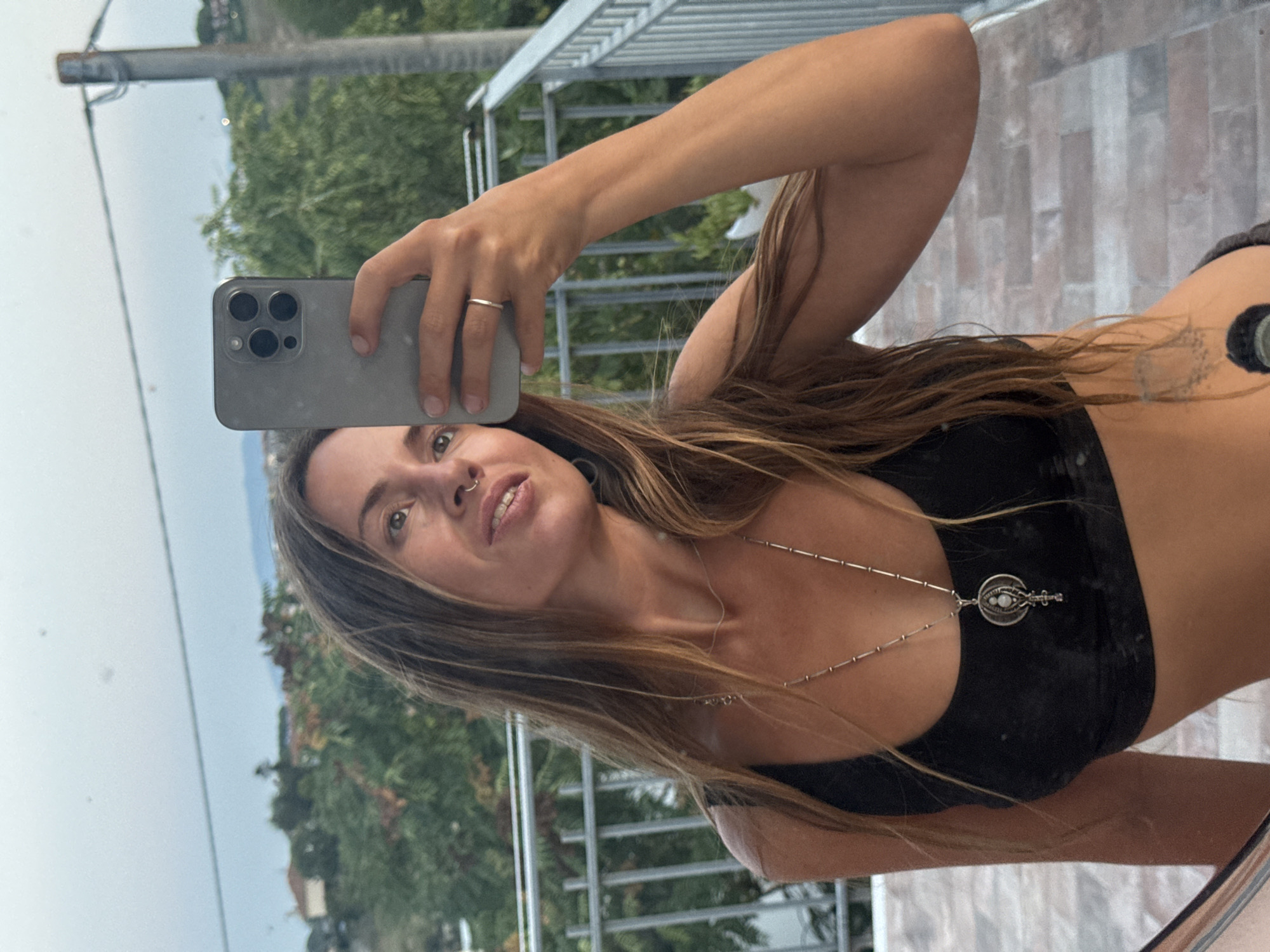 In a medium shot, a woman with long, light brown hair and a nose ring takes a selfie in what appears to be a balcony or patio area, reflecting in a mirror. She is wearing a black sports bra with a silver necklace featuring a circular pendant. Her right arm, holding a silver iPhone, is raised, and she wears a silver ring on her ring finger. The background shows greenery and a distant hillside with buildings. The overall lighting suggests it is daytime, possibly late afternoon or early evening.