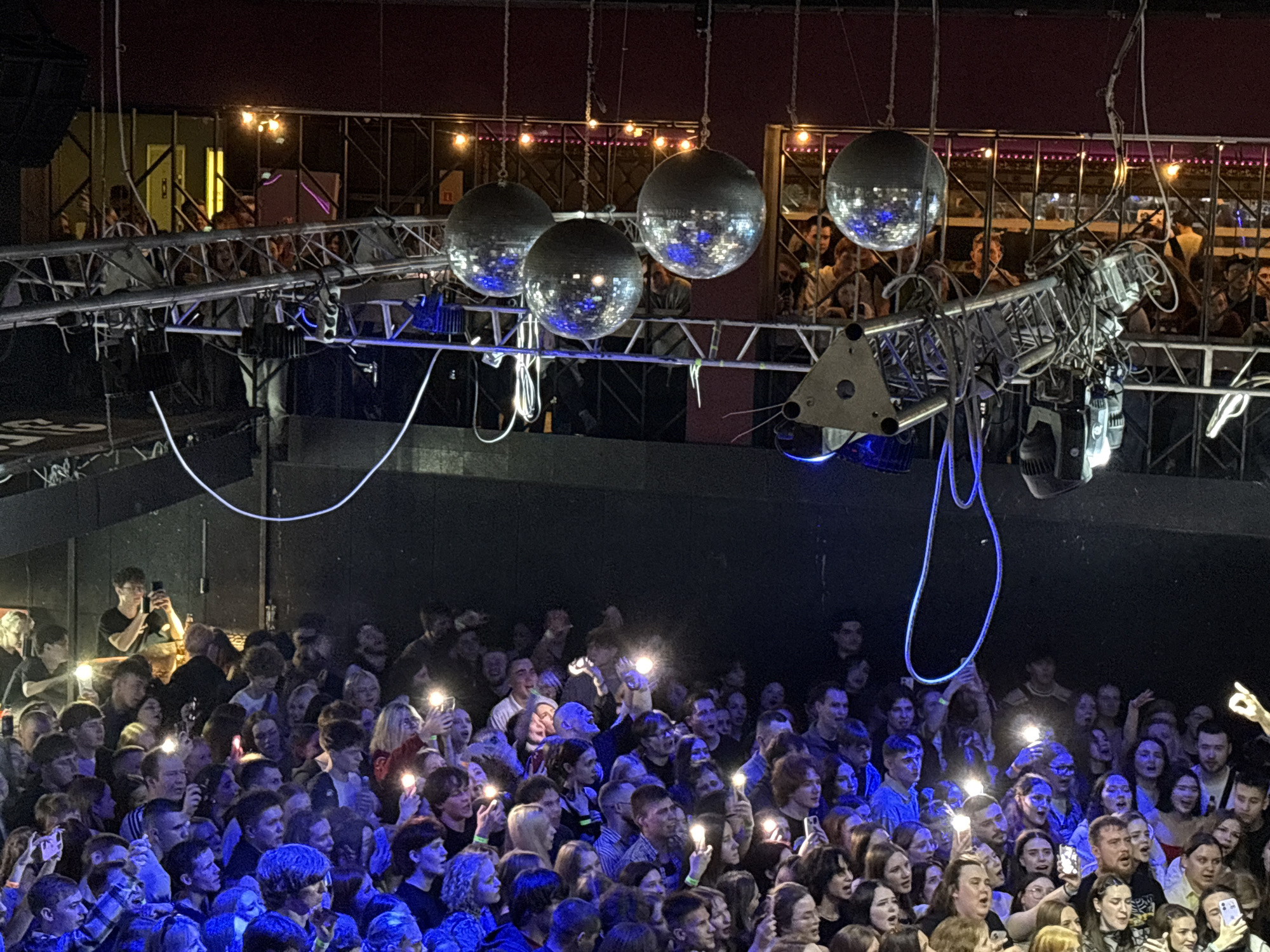 A large crowd of people, predominantly young adults, are gathered at a concert. The crowd is densely packed and lit from below with blue lights, with many individuals holding up their phones. A few individuals in the crowd have their phone lights illuminated. Overhead, hanging from the ceiling, are four mirrored disco balls suspended by chains. Behind the disco balls, there is a metal platform with people standing on it, looking down at the crowd. A metal scaffolding structure hangs from the ceiling containing lighting equipment, including blue spotlights. The location is most likely a concert venue based on the stage lighting and large crowd. Based on the request this image is in Izhevsk, Russia.