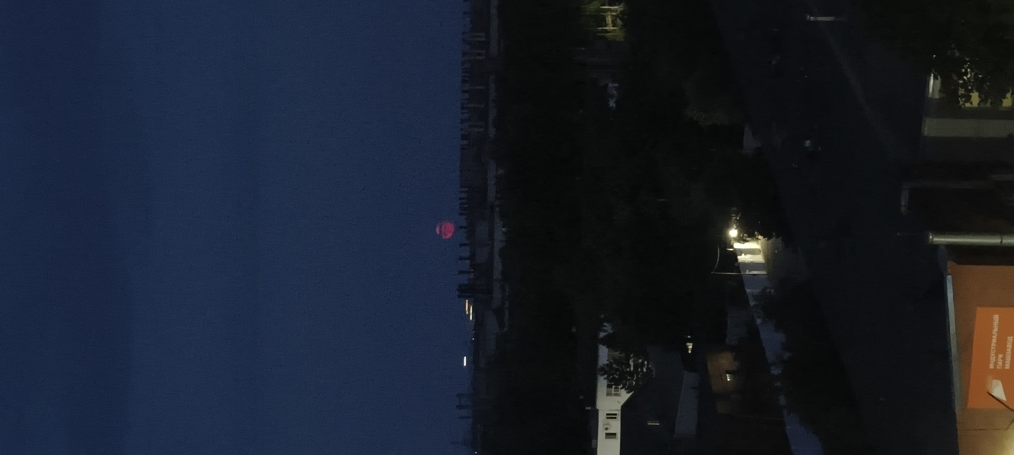 A view of a city skyline at dusk is captured, featuring a large, red moon in the upper portion of the frame. The moon appears slightly distorted and haloed. Below the moon, a silhouette of industrial buildings and structures extends across the horizon, with several tall, cylindrical smokestacks visible. The foreground of the image shows the dark silhouettes of trees and low-rise buildings, with some illuminated windows and a streetlamp casting a warm glow. In the bottom right corner, a partially visible orange structure with white lettering is present. The overall scene suggests an urban or industrial setting at twilight.