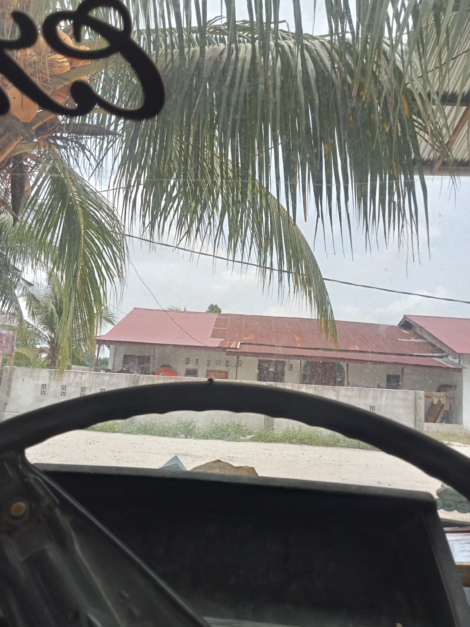 The image depicts a view from inside a vehicle, looking out through a windshield adorned with decorative lettering and palm fronds. In the foreground, the top of a steering wheel is visible. Beyond the windshield, a building with a red corrugated metal roof is the primary subject. The building appears to be a single-story structure with multiple windows and a wall in front of it. The surrounding environment consists of a sandy or dirt road, some greenery, and a cloudy sky. The image is likely taken in a tropical or subtropical region, as suggested by the palm trees. While the exact location of Melayu Besar Kota, Indonesia is not explicitly shown, the presence of tropical vegetation and architecture aligns with the general landscape of such areas. There are no individuals or direct human actions visible in the frame.