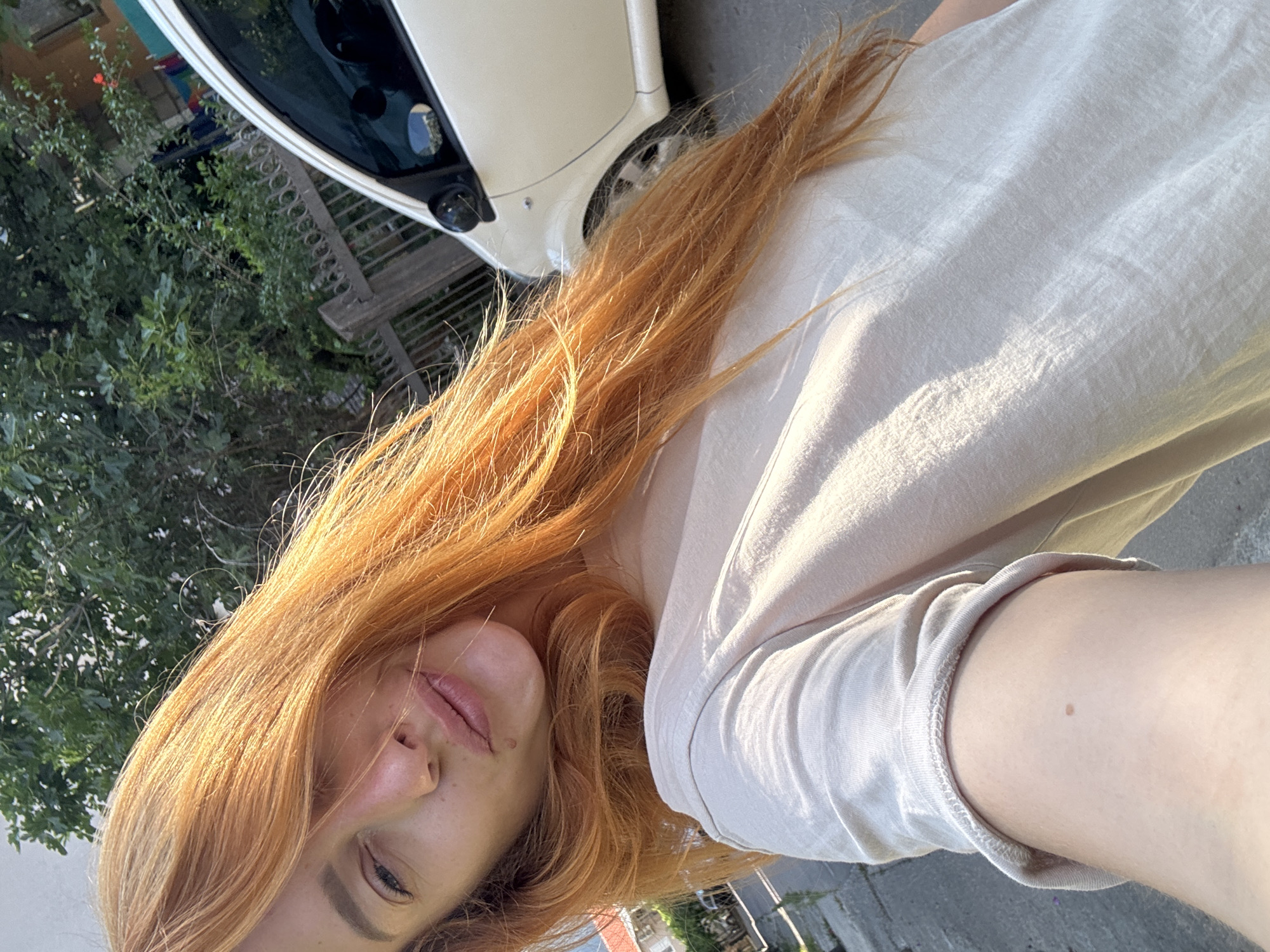 A selfie taken from a low angle shows a woman with vibrant orange hair in sunlight. She is wearing a light-colored t-shirt and is outdoors. In the background, a white car is parked near a fence and lush green trees, suggesting a residential or street setting within the city of Budva, Montenegro. Sunlight creates strong highlights on her hair and t-shirt, and the overall scene is bright and natural.