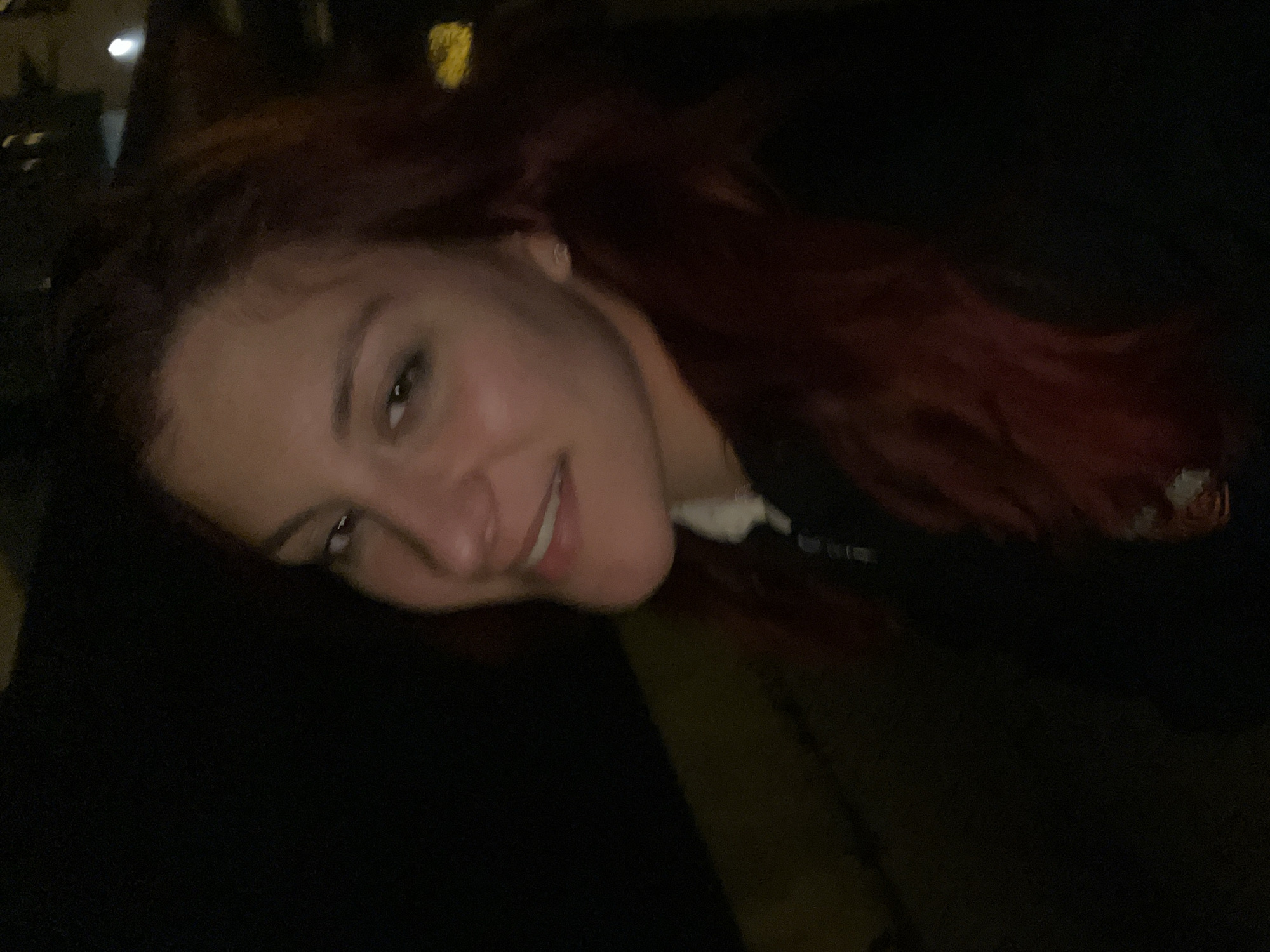 A person with reddish-brown hair is smiling and looking at the camera. The individual is wearing a dark-colored shirt and has fair skin. The environment appears dimly lit, and the location is not discernible from the image alone.