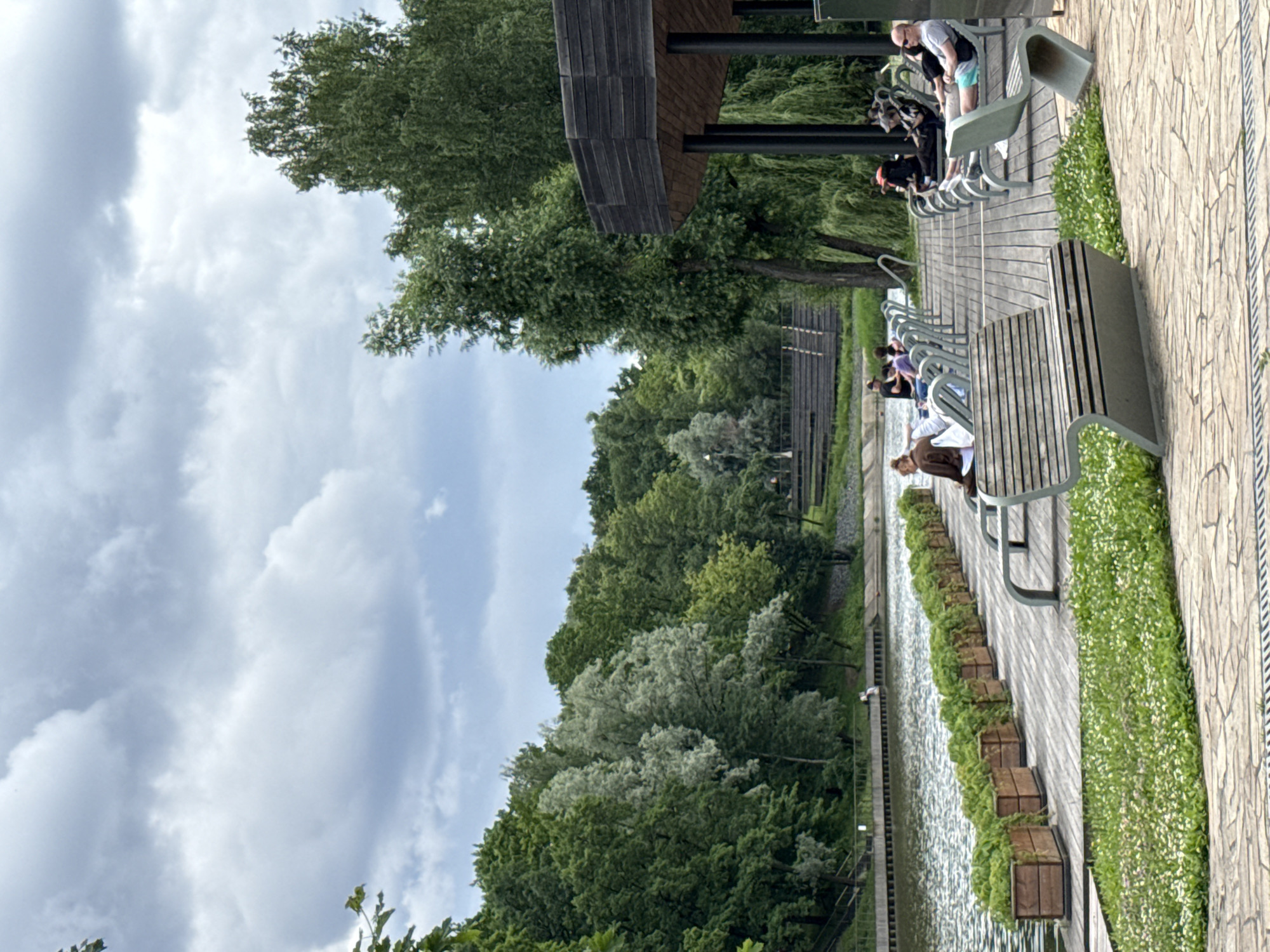 The image depicts a waterside promenade in Moscow, Russia, featuring a row of benches and planters along a wooden walkway. Several people are visible, some sitting on the benches, while others stand or walk along the path. The scene suggests a leisurely afternoon or early evening, with overcast skies indicating a cloudy day. The lush greenery of trees and plants bordering the walkway, along with the calm water, creates a serene park-like atmosphere.
The subjects include individuals of various ages, dressed in casual attire, enjoying the outdoor space. The objects consist of modern, minimalist benches and wooden planters filled with vibrant green foliage and small white flowers. The promenade itself is constructed with wooden planks, bordered by stone paving on one side. A contemporary wooden structure with dark supports, possibly a pavilion or canopy, is partially visible in the background, adding architectural interest.
The activity taking place is primarily passive: people are resting, conversing, or simply enjoying the scenery. There are no clear indications of a specific event, but rather a snapshot of everyday life in a public park. The weather appears mild, typical for a day with cloud cover, and the lighting is diffused. The presence of the water, trees, and the overall landscaping strongly suggests a public park or recreational area near a body of water. No visible text is present in the image.