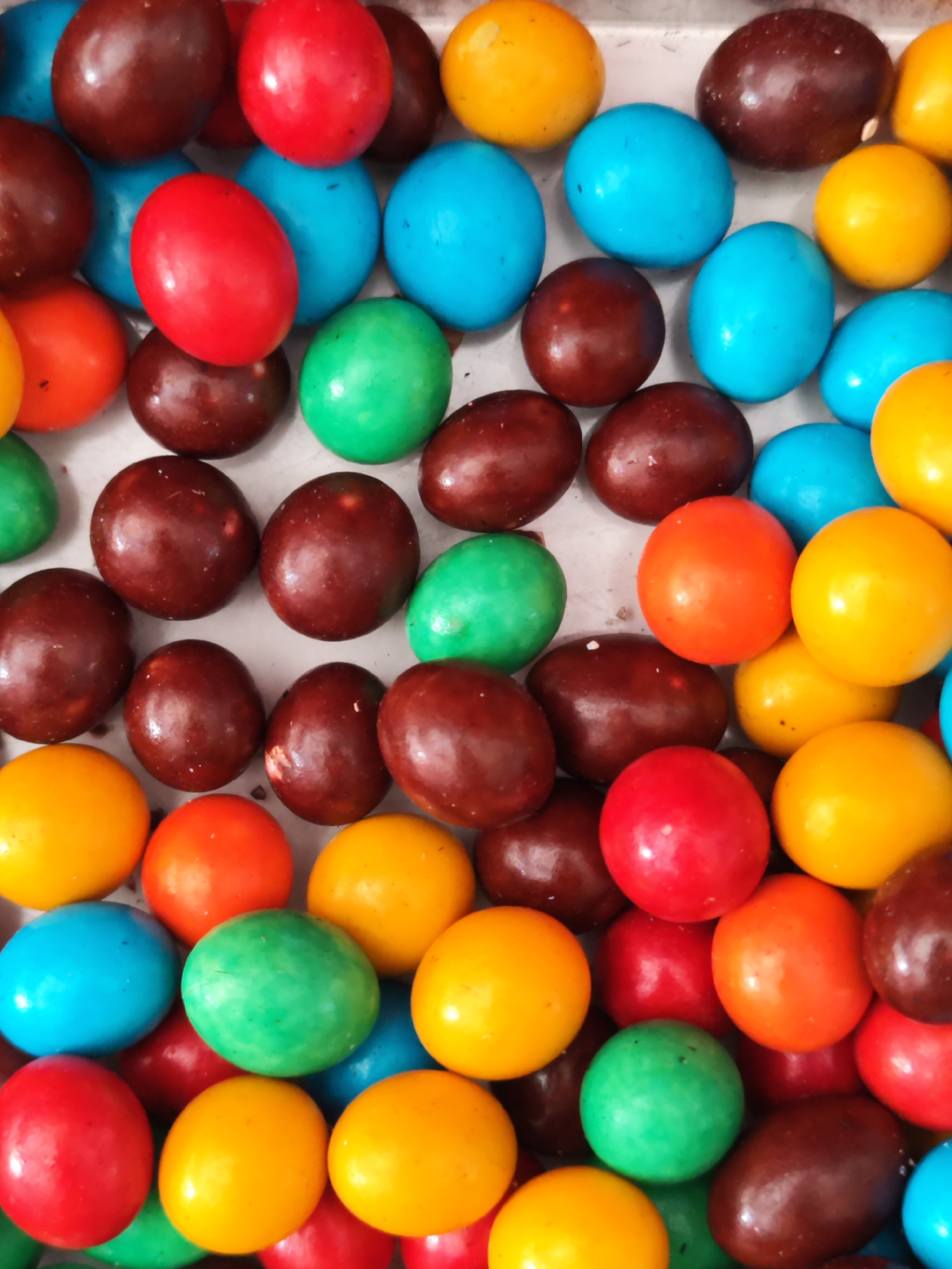 A close-up, overhead shot displays a chaotic jumble of brightly colored, oval-shaped candies. The candies are predominantly red, yellow, green, blue, and a dark chocolate brown. They are densely packed, with some overlapping and others nestled together. The surface they rest on appears to be a light-colored, possibly metallic, tray or surface, visible as white or very pale grey in the gaps between the candies. The lighting is even, casting no strong shadows, suggesting a diffuse light source or a flash. There are no people, animals, or any environmental context beyond the candies and the surface. The image is purely an arrangement of confectionery. No specific location within Sragen, Indonesia, can be determined from the visual information.