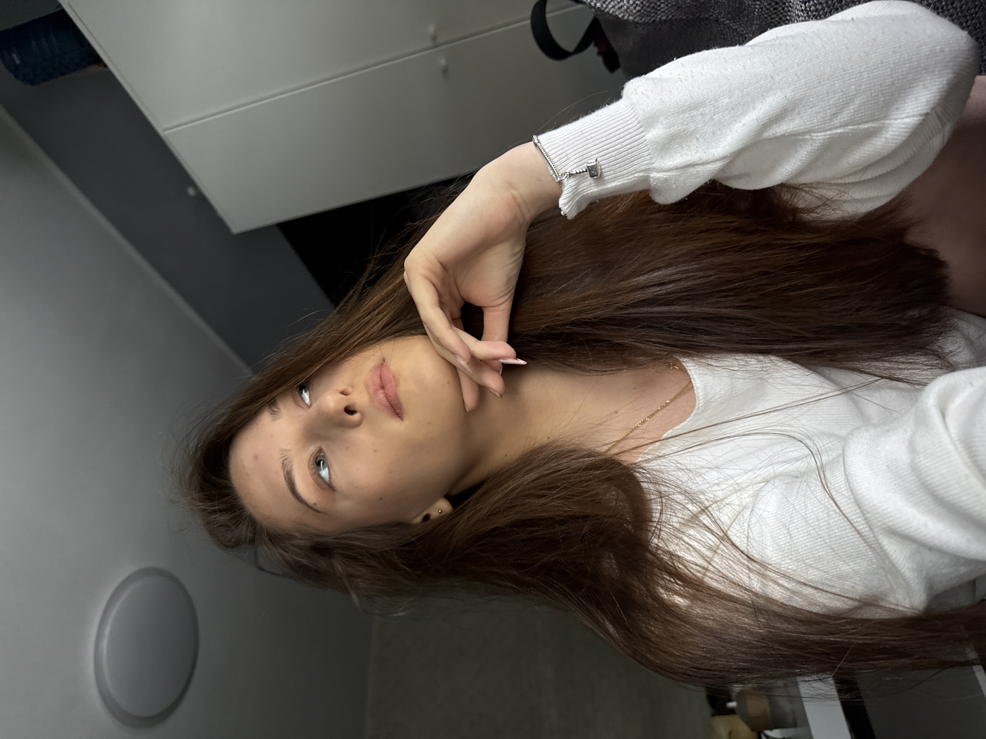 A young woman with long brown hair and fair skin is posing for a photograph. She is wearing a white long-sleeved shirt and a silver bracelet. Her left hand is raised to her face with her fingers gently touching her cheek. She is looking up and to the left with a thoughtful expression. The background consists of a white wall, a gray object that could be a light fixture or speaker, and what appears to be a section of a bed or couch with gray fabric. The setting is indoors. There is no indication that the image was taken in Sofia, Bulgaria.