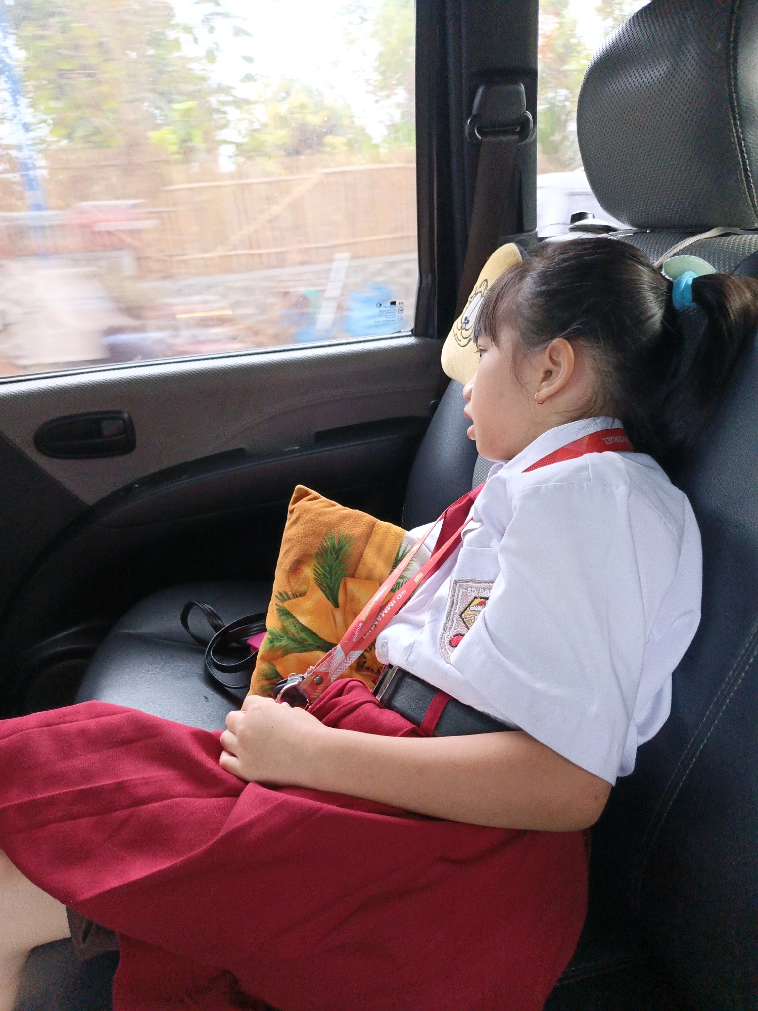 A young girl in a school uniform is sitting in the back seat of a car. She is wearing a white shirt with a red collar and a red skirt. A yellow and orange patterned pillow is tucked behind her. Her hair is tied back in a ponytail with a blue hair tie. The car is in motion, as suggested by the blurry view out the window, which shows trees and a wooden fence. The scene appears to be set during the daytime, with natural light filtering into the car. The girl's expression is neutral, and she is looking out the window. Visible text on a red lanyard reads "SD IMMANUEL". The location is Batu, Indonesia.