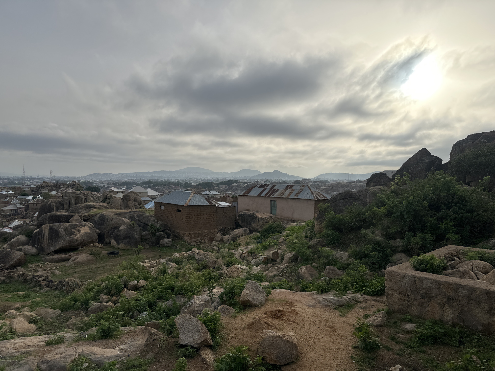 The image depicts a landscape in Jos, Nigeria, under a cloudy sky with the sun partially obscured. In the foreground, there is rough terrain with rocks, sand, and sparse vegetation. Two small, modest buildings with corrugated metal roofs are visible. In the background, a city skyline stretches into the distance, with hills or mountains further back. There are no people visible in the image.
