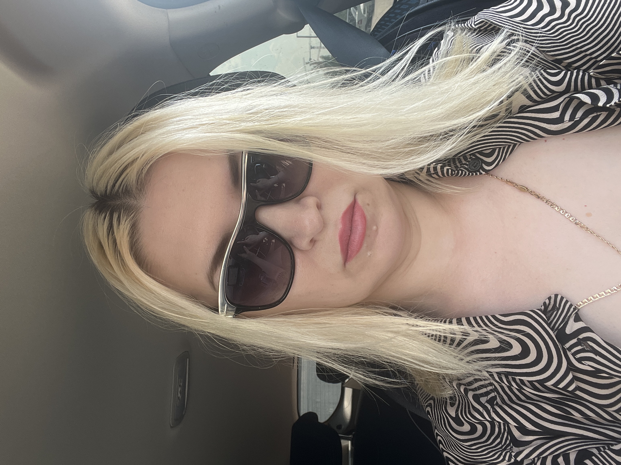 A close-up, selfie-style, horizontal shot features a blonde woman with fair skin and pink lipstick, wearing dark sunglasses with silver trim. Her hair is styled down, framing her face. She is inside a vehicle, indicated by the car's ceiling visible at the top of the frame and a seatbelt strap on the right. The woman is wearing a black and white patterned shirt. The scene appears to be outdoors, with a hint of a blurry background suggesting a city environment with buildings and vehicles. The photo does not contain any specific markers for the city of Tyumen, Russia.
