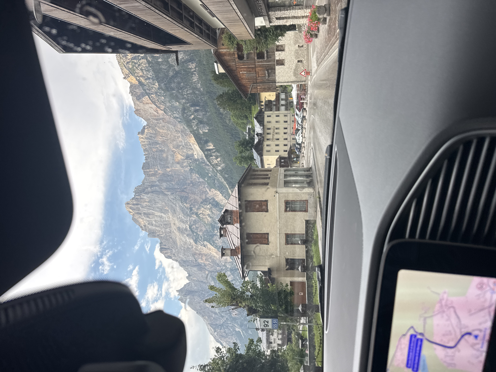 The image captures a view from inside a vehicle, looking forward through the windshield. A modern car's dashboard and navigation screen are visible in the foreground. The exterior scene shows a street in the town of San Vito di Cadore, Italy, with traditional alpine architecture. Mountains, covered in trees, dominate the background under a cloudy sky. Several buildings line the street, including a light-colored building with red shutters and a prominent red-tiled roof in the mid-ground. A road sign indicating "92 SS 51" is present. Cars are parked along the street, and some outdoor seating areas are visible. The street appears wet, suggesting recent rain. There are no people clearly visible in the image.