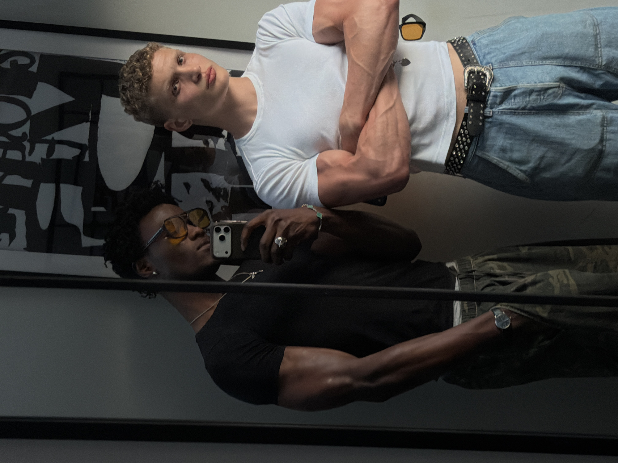 A reflection of two men is seen in a mirror. The man at the top of the frame is wearing a white t-shirt and blue jeans with a studded belt. He is looking down and towards the left. The other man is holding a mobile phone and is looking at his reflection, he is wearing a black t-shirt and camouflage trousers. He also has a watch on his wrist and sunglasses perched on his face. A poster is seen behind the man at the top of the image. It is not possible to determine the location of the image.
