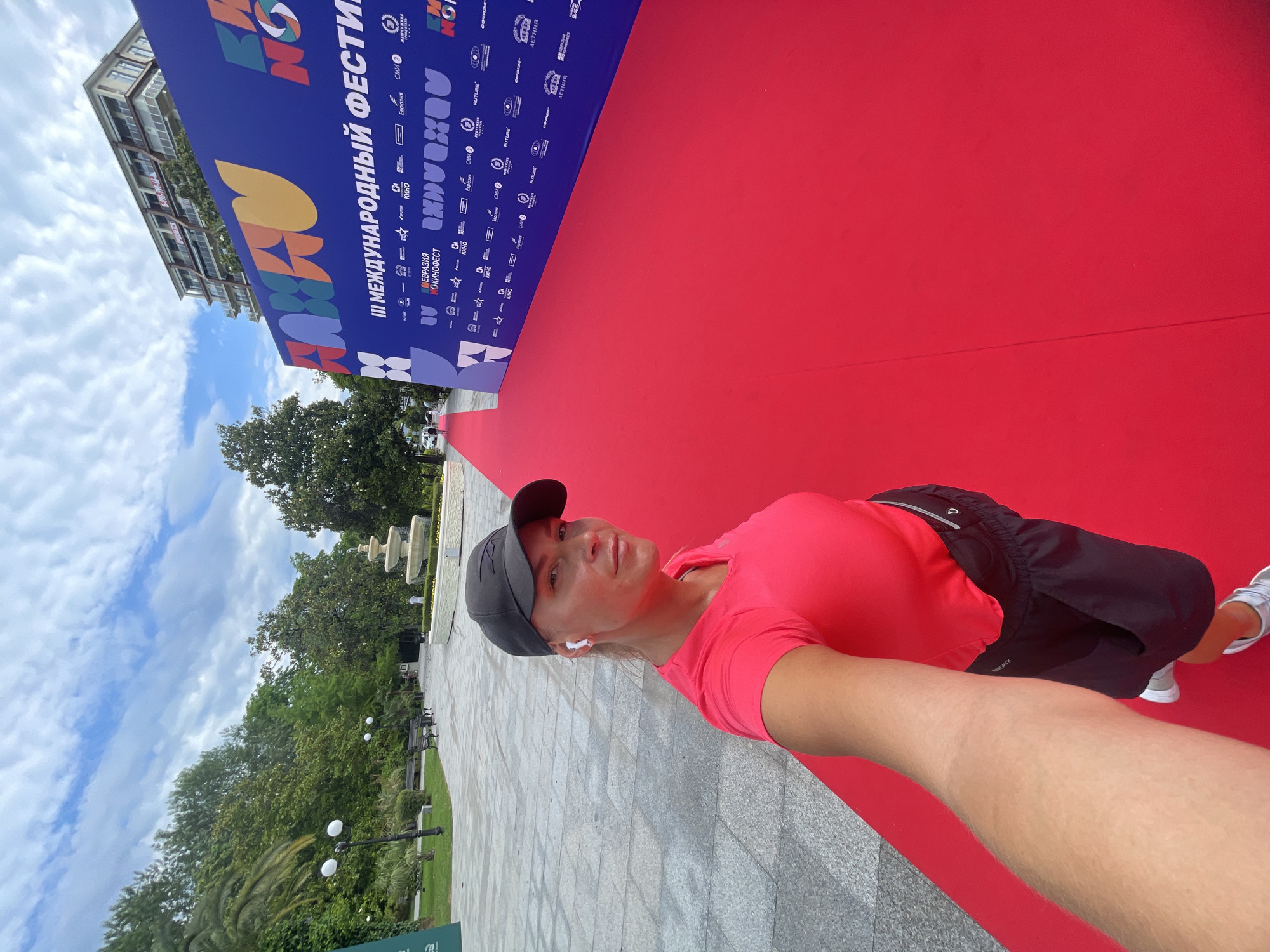 In this selfie, a woman with blonde hair, wearing a pink t-shirt, black shorts, a black baseball cap, and white earbuds, stands on a red carpet in an outdoor urban setting. The backdrop features a large blue banner with colorful graphics and text, including "III МЕЖДУНАРОДНЫЙ ФЕСТИВАЛЬ" (III INTERNATIONAL FESTIVAL). The banner is adorned with logos of various sponsors. To the left of the carpet and banner, a park area is visible with trees, benches, lampposts, and a tiered fountain. In the distance, a multi-story building with balconies can be seen. The paving stones in the foreground suggest a public walkway or plaza. The scene is consistent with an event held in the city of Sochi, Russia, likely at an outdoor venue.