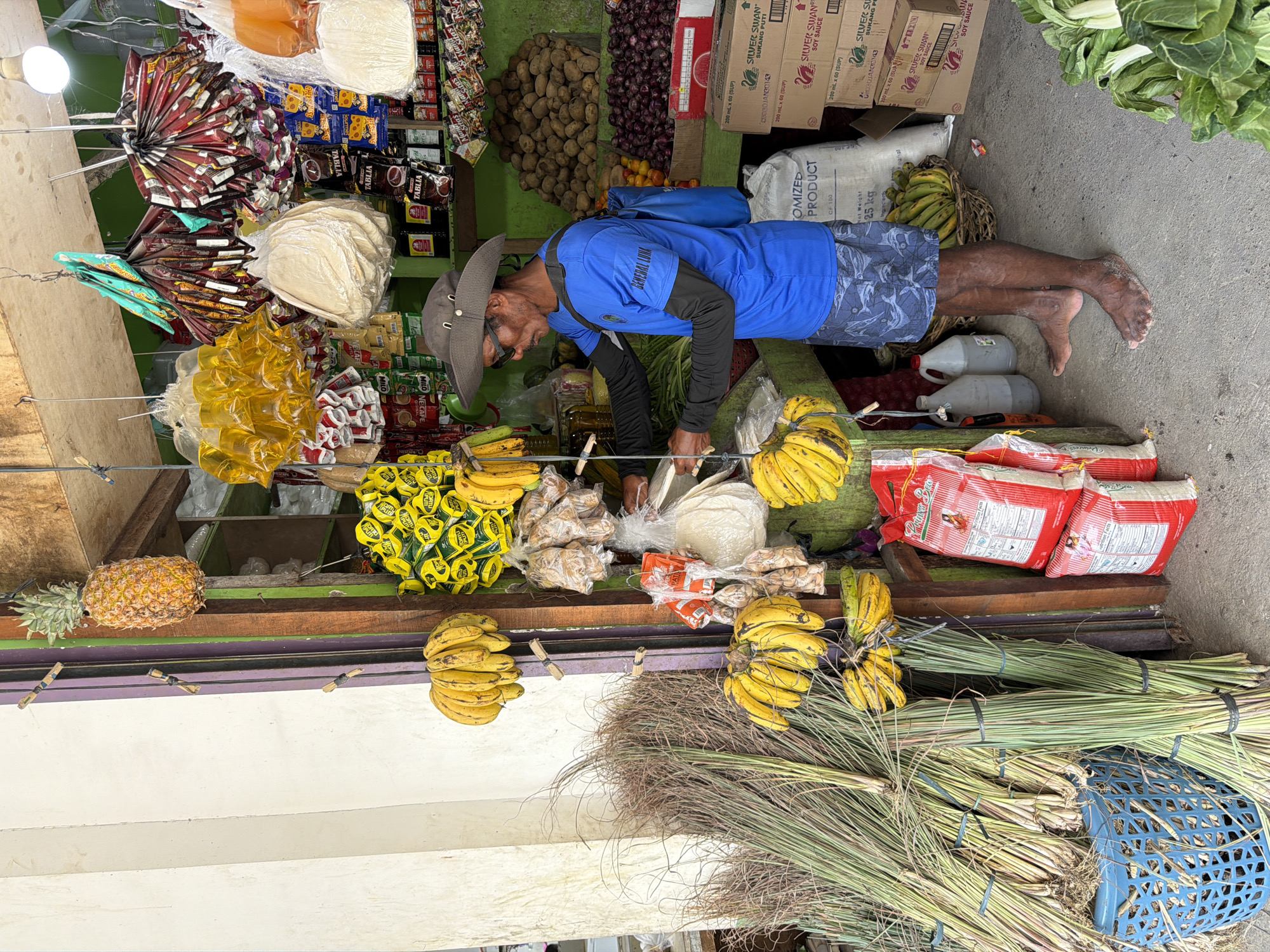 This image, rotated 90 degrees clockwise, captures a vibrant market stall scene in General Luna, Philippines, during daylight hours.
The central subject is a man, likely a vendor, standing barefoot on a concrete floor. He wears a grey wide-brimmed hat, dark sunglasses, a blue short-sleeved shirt with long black sleeves underneath, and patterned blue and white shorts. A blue bag is slung over his shoulder, and a discernible text on his shirt reads "BEMERAL LUNA", aligning with the provided location. He appears to be arranging or preparing goods at a wooden counter.
The stall is abundant with various produce and packaged items. To the man's left, large bunches of bright yellow bananas hang, some tied together. Further left, a pineapple rests on a wooden shelf. Below the counter, more bananas are visible. To his right, bundles of what appear to be dried grass or leaves are piled up next to a blue plastic basket, with two red sacks visible behind them.
The shelves behind the man are packed with groceries. There are large bags of potatoes, reddish-purple onions, and what looks like green leafy vegetables. Numerous packaged goods include boxes of "SILVER SWAN SUKANG PUTI" (vinegar) and "SILVER SWAN SOY SAUCE," some indicating "200 mL x 60 (SUP)". Other items include bags labeled "OMIZED PRODUCT 25 kg", possibly rice or flour, and red bags with "Princess" written on them, likely another brand of rice. Above the counter, there are clear plastic bags of flatbreads or crackers, yellow pouches of cooking oil, and packaged drinks like "MILO" and "NESCAFE". Dark packets labeled "TABLIA" are also visible. A single exposed light bulb illuminates the upper section of the stall.
The scene suggests a lively local market, showcasing fresh produce alongside essential household items and packaged foods, characteristic of daily commerce in the Philippines.
