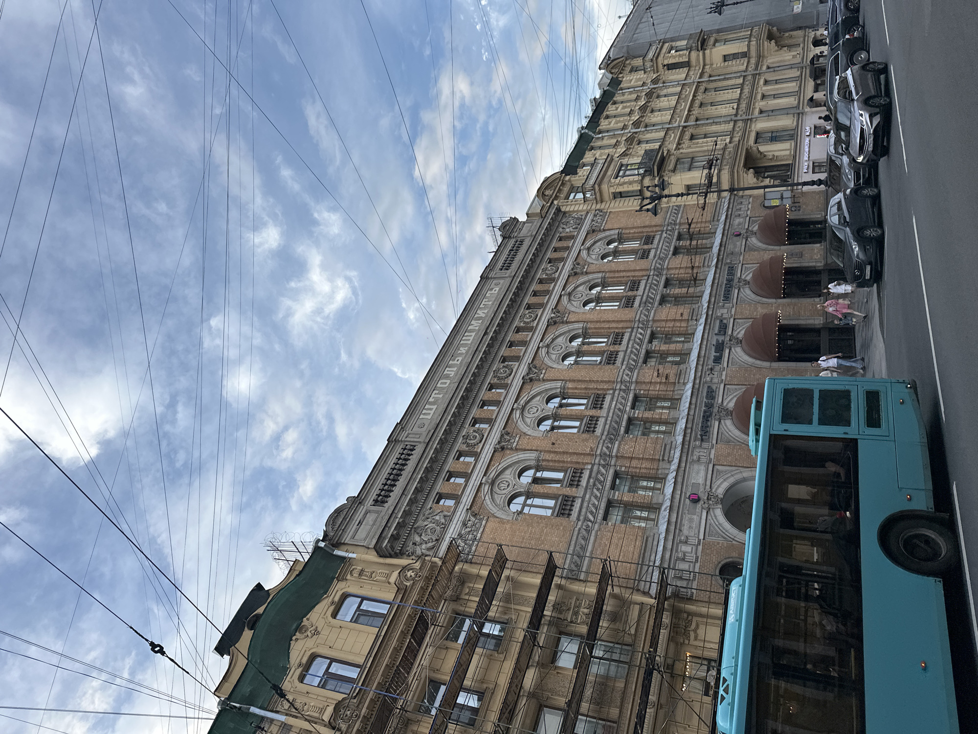 The image depicts a street scene in Saint Petersburg, Russia, likely on Nevsky Prospekt, given the architectural style and the presence of trolleybus wires. In the foreground, a bright turquoise trolleybus is traveling on the right side of the road. Behind the trolleybus, several cars are parked or moving, including a dark gray SUV, a silver sedan, and other vehicles. The street is paved, and white lines delineate the lanes.
The background is dominated by a row of historic buildings with intricate facades. The building directly behind the trolleybus is an ornate brick structure with multiple arched windows and decorative elements. Above the arched windows, there is text in Cyrillic, which appears to read "ШТОЛЬН" and "ШМИТЬ". To the left of this building, another building is partially covered by scaffolding, suggesting ongoing renovation or construction. This building is lighter in color and also features detailed architectural ornamentation. Further to the right, the facade of another yellow building with balconies and multiple windows is visible, characteristic of St. Petersburg's classical architecture. The sky above is partly cloudy with a mix of blue sky and white clouds, and numerous electrical wires crisscross the frame. A few pedestrians are visible on the sidewalk in front of the buildings, walking and interacting. The overall atmosphere suggests a bustling urban environment in St. Petersburg.