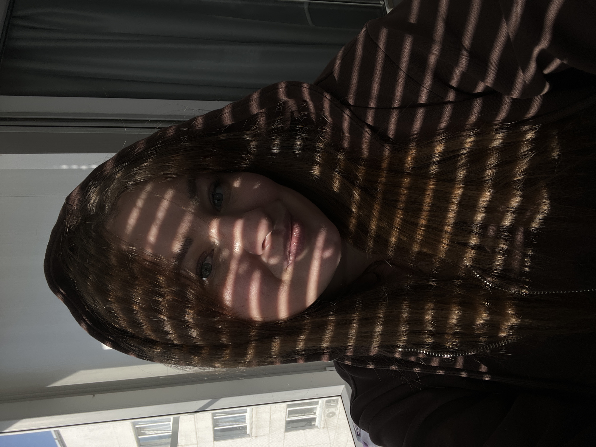 A photograph shows a woman's face, partially obscured by the shadows of window blinds. She is wearing a brown hooded sweatshirt. The background shows buildings across the street with a clear sky visible. The location is likely Chaikovsky, Russia.