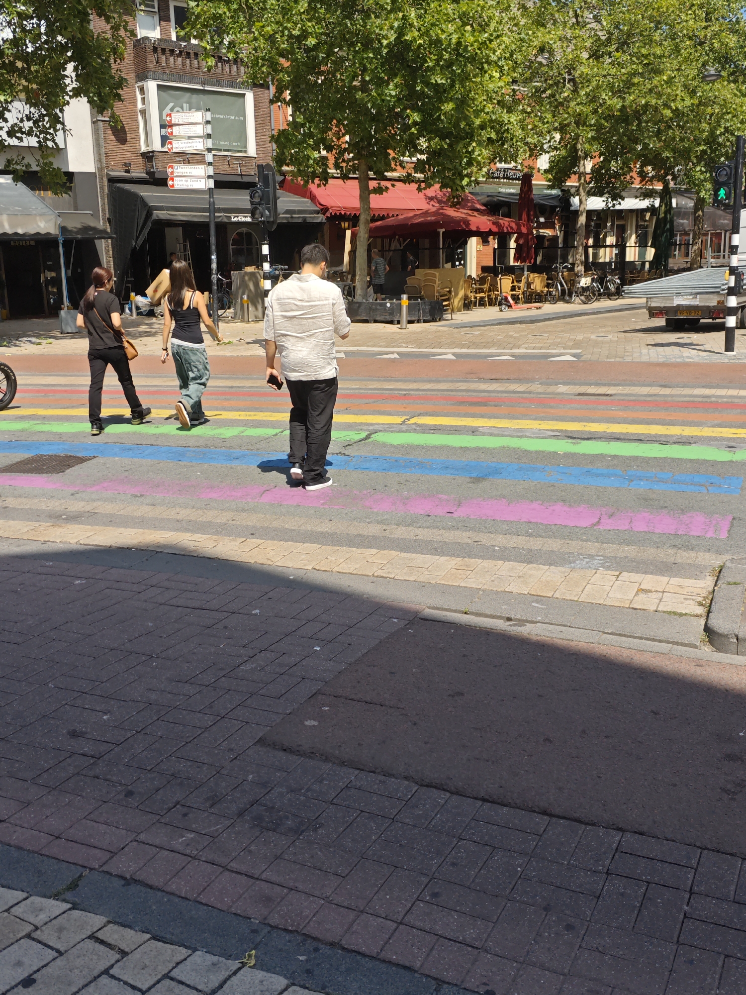 The image shows a street scene in the city of Tilburg, Netherlands, with a rainbow-colored crosswalk in the foreground. Three individuals are walking across the crosswalk. On the left, a person with dark hair wearing a black t-shirt and dark pants walks away from the camera, carrying a tan purse over their left shoulder. In the middle, a person with dark hair wearing a black tank top and light-colored, baggy pants also walks away from the camera. To the right, a person wearing a light-colored, striped short-sleeved shirt and dark pants walks away from the camera, holding a smartphone in their right hand.
In the background, there are several buildings, some with awnings and outdoor seating areas for cafes. Trees with green foliage line the street. Traffic lights are visible, with a green light illuminated on the right side of the image. A trailer is parked on the right side of the street. The ground is paved with cobblestones and asphalt. The overall scene suggests a sunny day in a commercial area of the city.