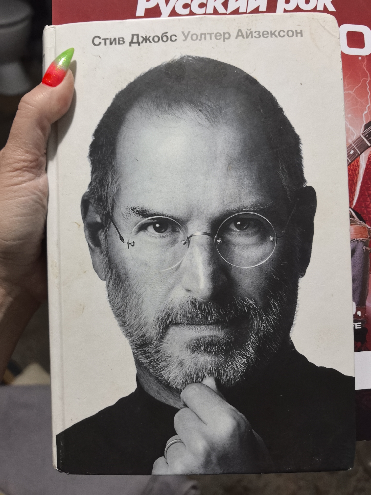 A person's hand, adorned with long, pointed nails featuring a vibrant red-to-neon-green gradient, holds a book. The book prominently displays a black and white portrait of Steve Jobs on its cover. Jobs is depicted with a beard and round, rimless glasses, gazing forward with a contemplative expression, his left hand gently resting on his chin. Above his image, text in Cyrillic reads "Стив Джобс Уолтер Айзексон," indicating the book is the Russian translation of Walter Isaacson's biography of Steve Jobs.
Partially visible behind this book is another book with a red and white cover. The title, "РУССКИЙ РОК" (Russian Rock), is visible at the top, and elements like a guitar and lightning bolts are discernible on its cover. The background is softly blurred, showing indistinct indoor elements, including what appears to be a ceramic fixture and a plain greyish-purple surface. The context of the Russian language text on both books, particularly "РУССКИЙ РОК," aligns with the specified location context of Noviy, Russia. The scene captures a moment of holding or presenting books in an indoor setting.