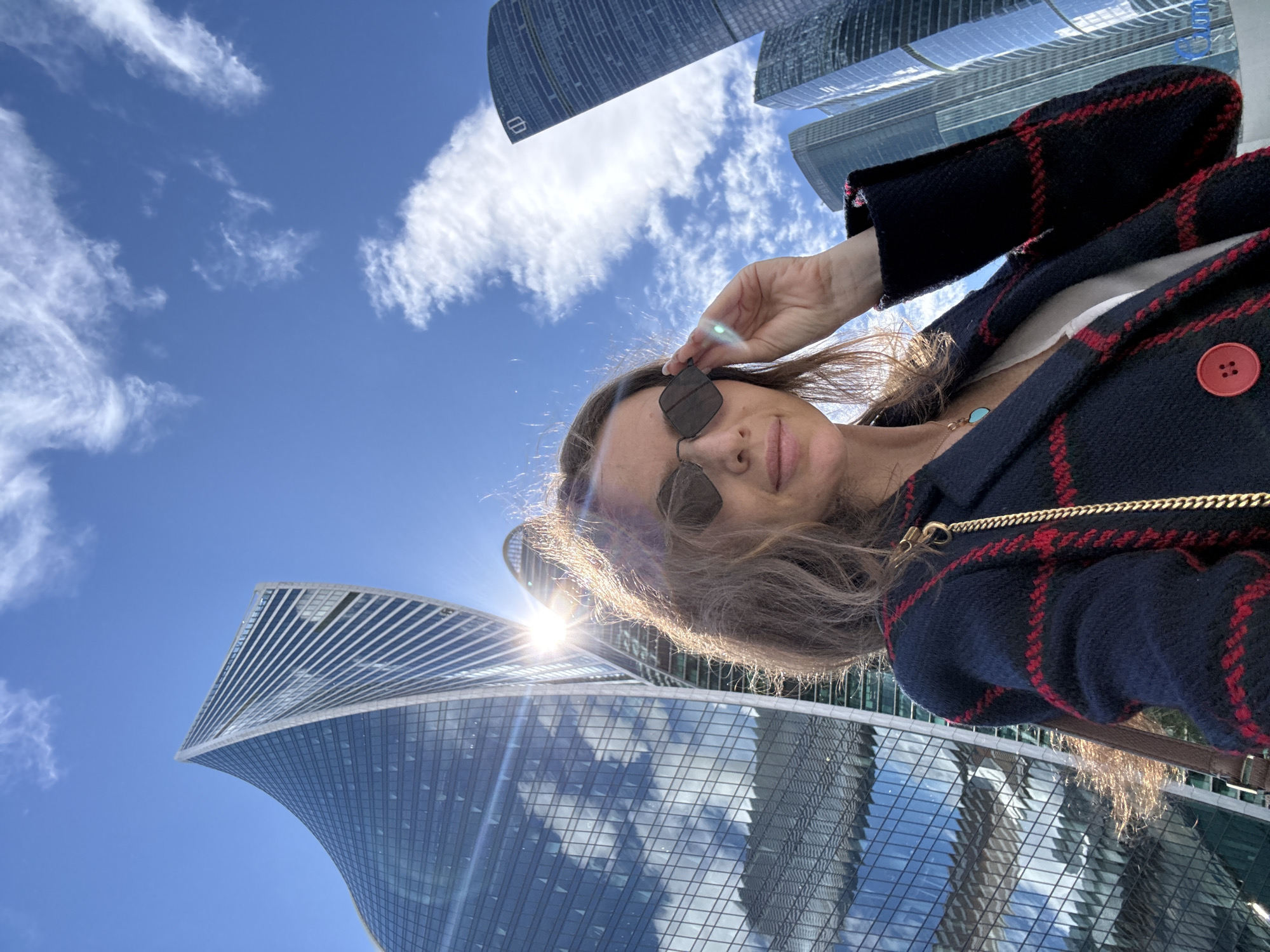 A medium shot captures a woman from the chest up, posing for a selfie in front of a backdrop of modern skyscrapers in Moscow, Russia. The woman, with long, wavy brown hair and wearing sunglasses, is dressed in a dark blue blazer with red trim and a red button. She is adjusting her sunglasses with her right hand. The sun is shining brightly, creating a lens flare that casts a golden glow on her hair and face. The skyscrapers, with their sleek, glass facades, reflect the blue sky and scattered white clouds. The buildings appear to be part of the Moscow International Business Center, commonly known as Moscow-City, a commercial district. The architecture is diverse, with some buildings featuring distinctive twisting designs. The overall scene conveys a sense of urban sophistication and a bright, sunny day.
