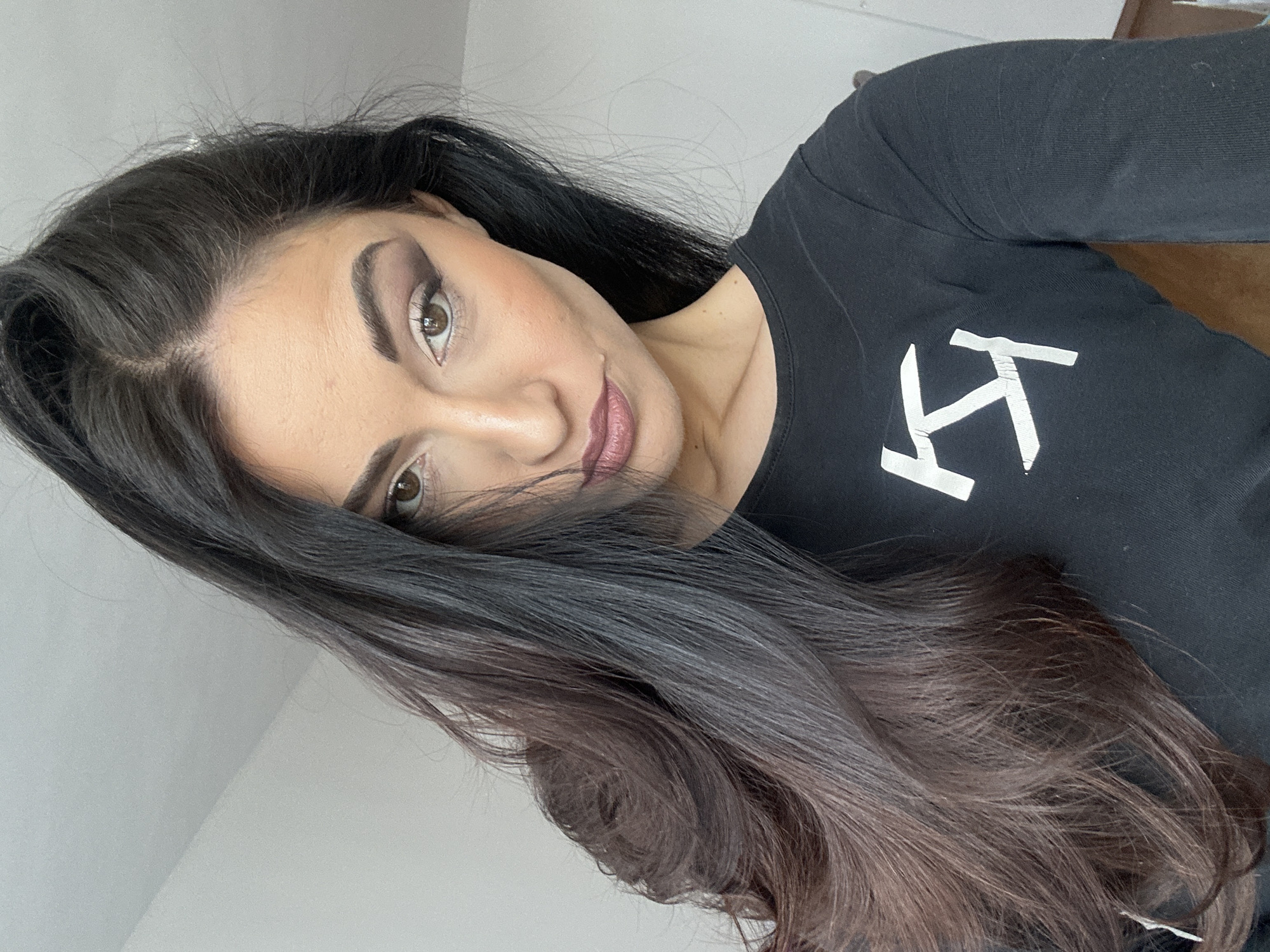 The image displays a close-up portrait of a woman, taken at a tilted angle. She has long, dark brown hair with lighter, auburn-toned ends, styled to cascade over her shoulder and partially obscure the right side of her face. Her dark brown eyes are adorned with smokey eyeshadow and eyeliner, complementing her well-defined, dark eyebrows. She wears a muted plum-colored lipstick and subtle blush. Her expression is neutral, and she looks directly at the camera.
She is wearing a black long-sleeved top that features a prominent white, stylized logo on the chest. The logo consists of two intertwined or mirrored block letters, resembling "KI" or "KT". The background is a plain, light-colored wall, providing a simple setting. The lighting is soft and even, characteristic of an indoor environment.