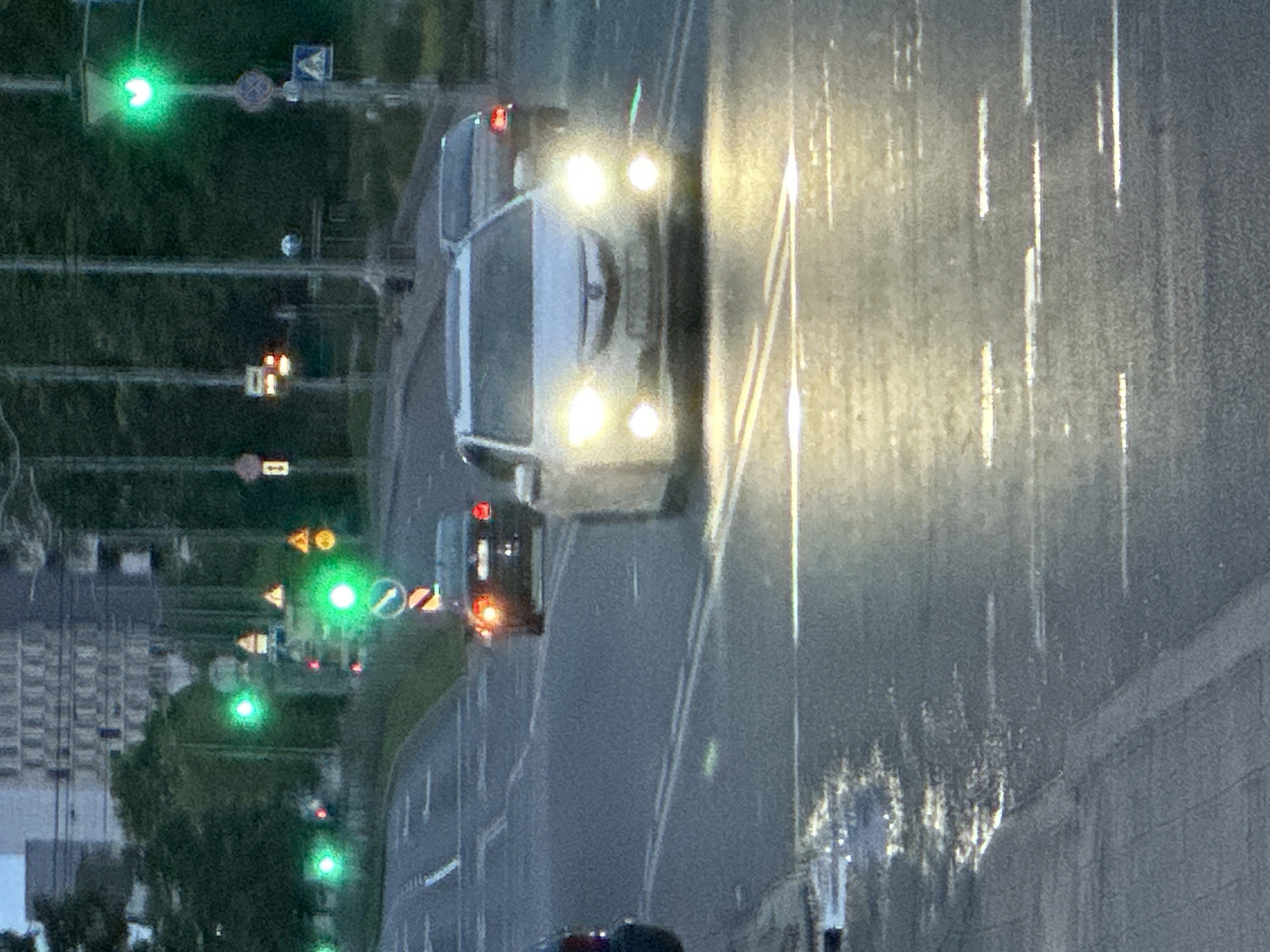 The image depicts an urban road scene during twilight hours in Minsk, Belarus. The asphalt road is wet, showing reflections from vehicle headlights and overhead lights.
In the foreground, a white SUV, identified as an Acura RDX, faces the viewer with its headlights and fog lights actively illuminated. Its front license plate, partially obscured and blurred, reads `7886 IB-7`. To the left, the rear of a dark-colored vehicle is partially visible. Further down the multi-lane road, a dark sedan, possibly a BMW, is seen from the rear with its red taillights illuminated, positioned under a set of traffic lights.
Several traffic lights are active and displaying green signals. One prominent green traffic light on the right features a unique heart-shaped signal within its display, characteristic of Minsk. Another traffic light, above the dark sedan, also shows a green signal, possibly an arrow.
Multiple road signs are visible on poles alongside the road. These include yellow triangular warning signs with black symbols, a round blue mandatory turn sign with a white arrow, and a round blue 'no stopping' sign with a diagonal red line. A square white sign with a red border and the number '1' inside is also present on a pole to the left.
The road features visible white dashed and solid line markings. Trees and dense foliage line the sides of the road. In the background, blurred multi-story buildings are visible, consistent with an urban environment. Utility poles or lamp posts are spaced along the road, supporting traffic signals and signs. No pedestrians are visible in the frame.