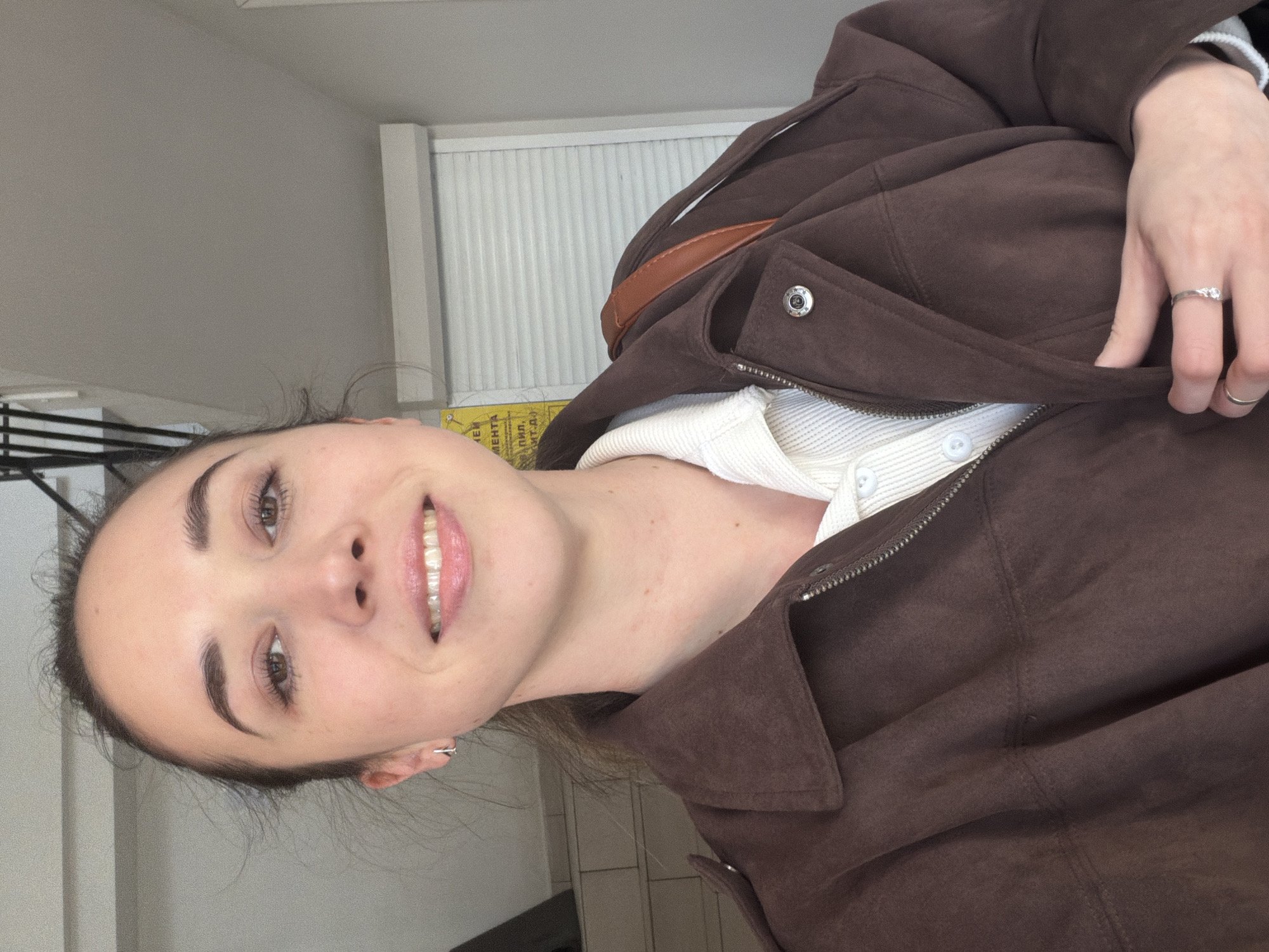 A woman is pictured indoors. She is wearing a brown suede jacket over a white button-up shirt. She is also wearing rings on her fingers. The background includes a white wall and a window with white blinds. A sign in Russian is partially visible. Without additional information, the precise location within Rostov-on-Don, Russia, cannot be determined.