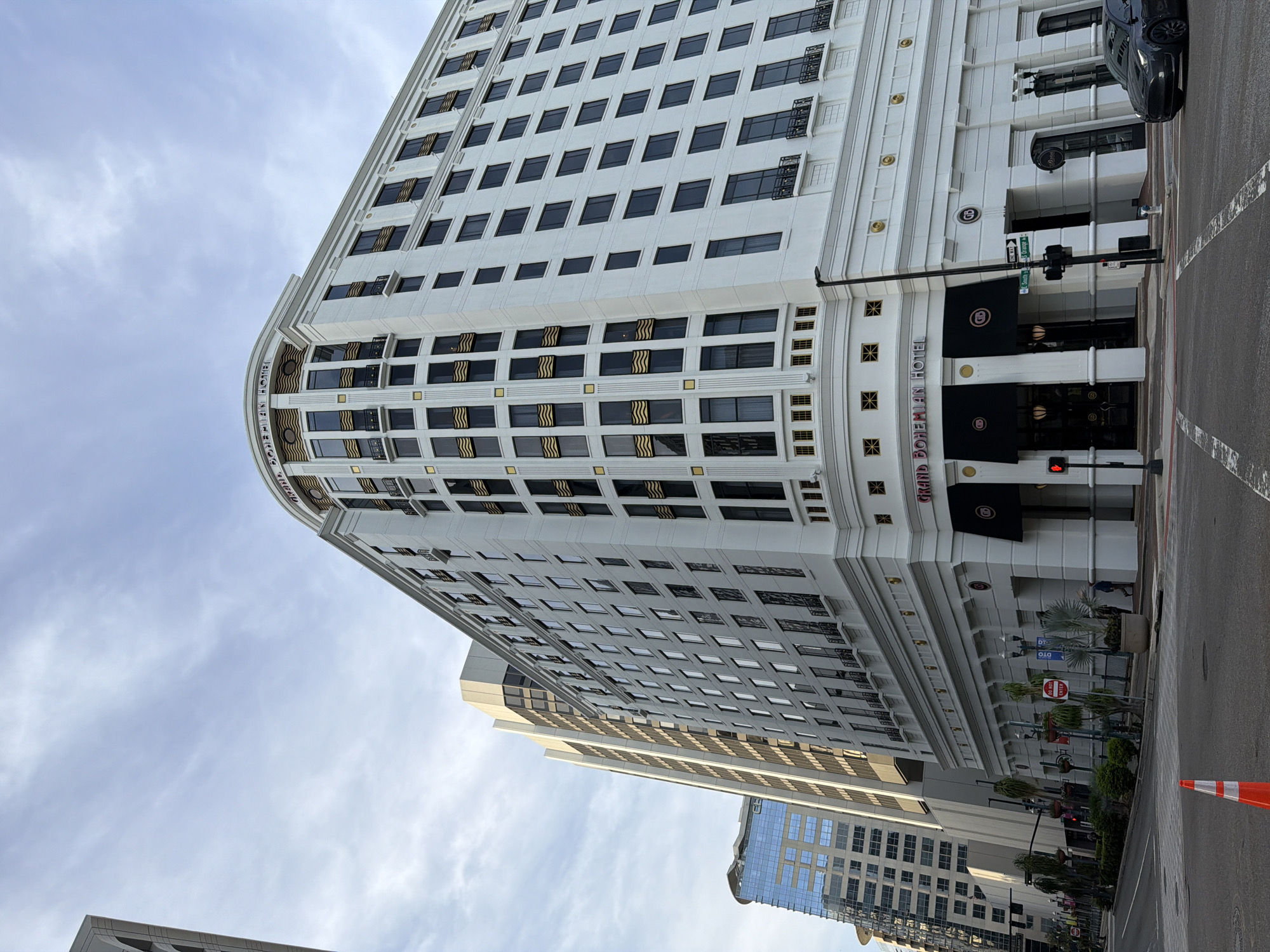 The image shows a low-angle, exterior view of the Grand Bohemian Hotel in Orlando, Florida, United States of America. The hotel is a large, white, multi-story building with many windows and decorative gold accents. The building has a curved corner at the top, which features the hotel's name in red lettering. Below this, there are black awnings over storefront entrances. The hotel is situated in a city environment, with other tall buildings visible in the background. A street runs in front of the hotel, with traffic lights, street signs, and a parked car. An orange traffic cone is visible in the foreground on the street. The sky is overcast with clouds.