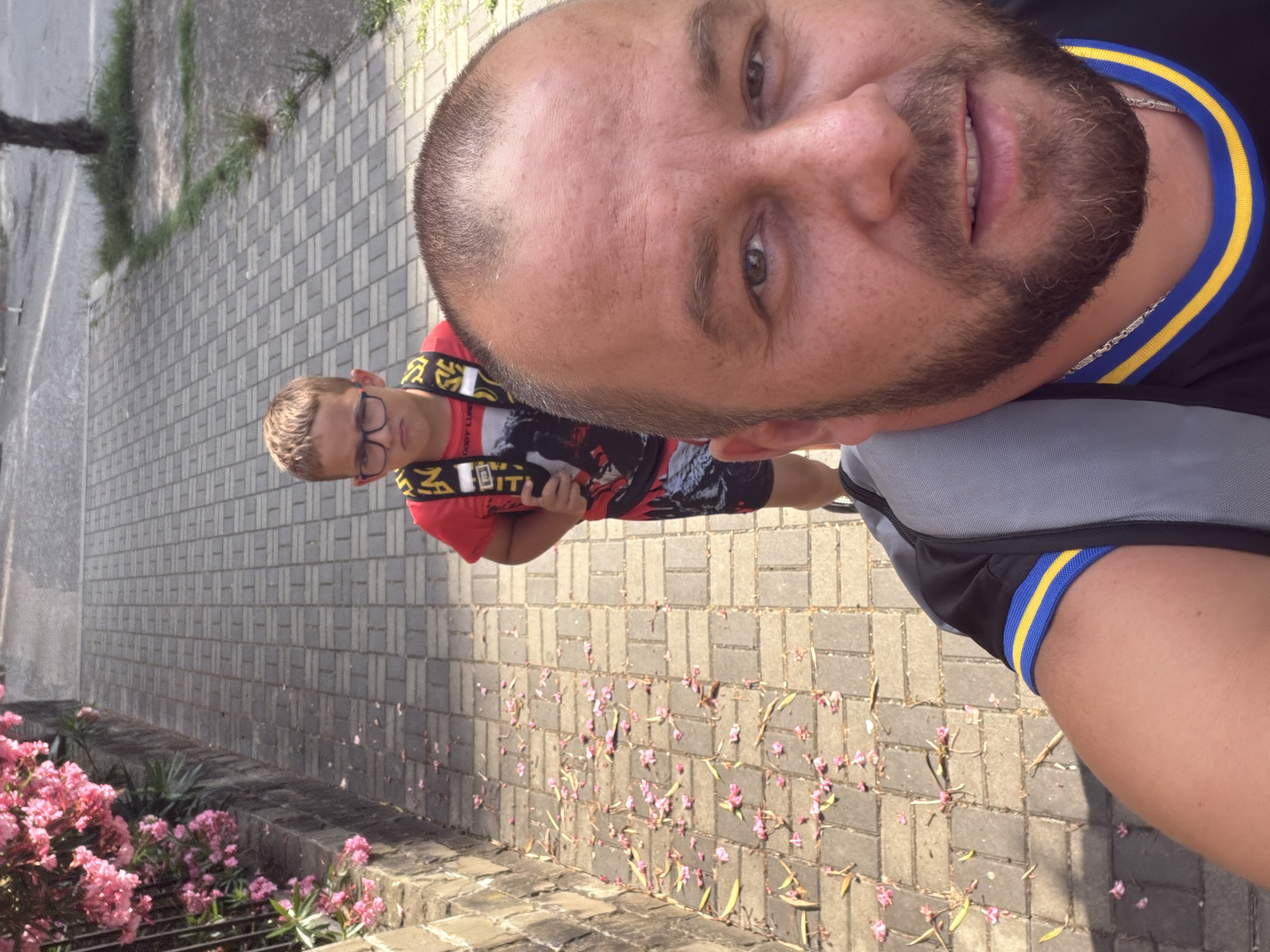 The image is a selfie taken outdoors, possibly in Sochi, Russia, given the context. The foreground shows the face of an adult male, who appears to be taking the photo with a wide smile. He has short, dark hair, a beard, and is wearing a black t-shirt with a blue and yellow striped collar. He is also wearing a grey bag. To his left, a young boy is visible. He has short, light brown hair and is wearing glasses. He is wearing a red t-shirt with a black and white graphic design and what appears to be a yellow and black lanyard or strap around his neck.
The background of the image shows a paved walkway with interlocking brick patterns. Scattered across the walkway are small pink flower petals and fallen leaves. To the left of the walkway, there are some green plants and grass. Further in the background, a building with a tiled wall can be seen. The lighting suggests it is daytime, and the presence of fallen petals and leaves might indicate a spring or autumn season, or a time when flowers are in bloom and shedding. The overall mood appears casual and relaxed. There is some visible text on the boy's t-shirt, but it is not clearly legible.
