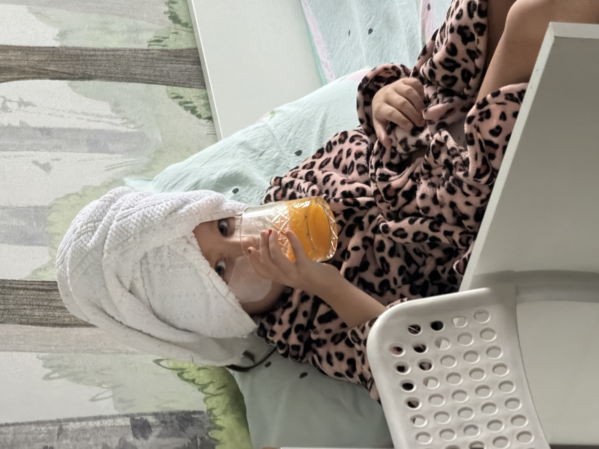 A young child, likely a girl, is lying down in what appears to be a bed, possibly in an indoor setting. She is wearing a white towel on her head and a pink and black animal print robe. The child is holding and drinking from a glass or small pitcher containing an orange liquid, presumably juice. She is also wearing a white mask on her face. A white plastic chair is visible near her. The backdrop includes a wall with a nature-themed design. It is impossible to determine from the image if this image was taken in Krasnoyarsk, Russia, or to know the subject's demographics beyond the visible physical appearance.