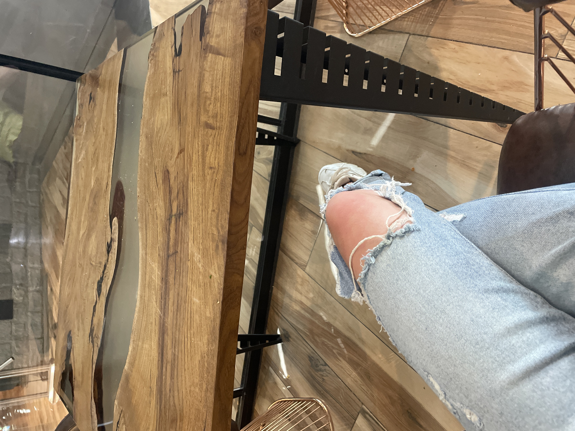 This image captures an angled, close-up view of an indoor setting, likely in Sofia, Bulgaria, focusing on a person's legs and a modern table. The perspective suggests the viewer is seated.
The main subject is a person's legs, clad in light-wash blue jeans that are heavily distressed with large rips at the knees, exposing skin. They are wearing white sneakers with visible laces and a chunky sole.
Dominating the left side of the frame is a unique table featuring live-edge wooden slabs joined by a clear epoxy resin 'river', allowing a partial view of the floor beneath. The wood displays prominent grain patterns and natural imperfections. The table is supported by a black metal frame, which includes a horizontal bar designed with a series of rectangular cutouts, giving it a contemporary, possibly industrial aesthetic.
The floor is covered with light-toned wooden planks arranged in a chevron or herringbone pattern, contributing to the stylish interior. In the lower right background, a glimpse of a dark brown, possibly leather, chair or sofa is visible. Another metallic, wireframe object, possibly a decorative basket or part of a furniture piece, can be seen on the floor.
The lighting indicates an indoor daytime environment. There is no visible text in the image.