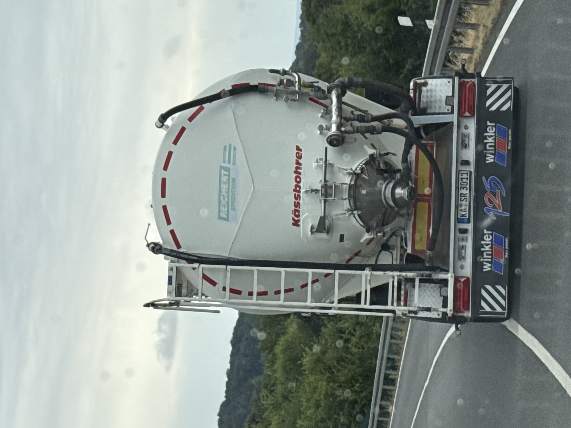 A white tanker truck is driving on a road with trees and hills in the background. The truck has a ladder on the side and a logo that says "REICHERT". The back of the truck has a license plate that says "KA SR 3011" and a logo that says "winkler". The truck is in motion. The sky is cloudy.