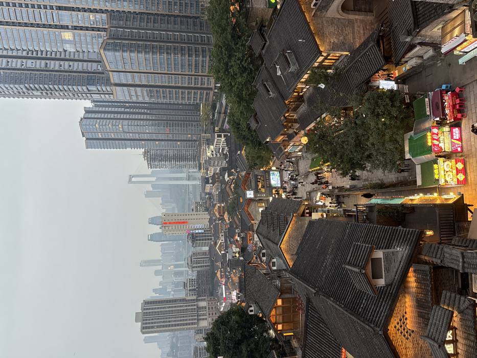 The image depicts an elevated view of an urban landscape at dusk or early evening in Yuzhong District, China. The scene is characterized by a blend of traditional architecture in the foreground and a modern skyscraper skyline in the background.
In the immediate foreground and midground, a complex of traditional-style Chinese buildings with multi-layered dark grey tiled roofs is visible. These structures feature illuminated windows and doorways, indicating active businesses or residences. A winding pedestrian street courses through these buildings, densely populated with numerous individuals walking in various directions. Several brightly lit vendor stalls, predominantly green, red, and yellow, are clustered at the lower right of the street, displaying Chinese characters on their signage and attracting pedestrians. Warm artificial lighting emanates from the buildings, streetlights, and vendor stalls. Green foliage from trees is interspersed among the buildings.
The background presents a vast array of modern skyscrapers, tall and multi-storied, constructed from glass and concrete, with some internal lights visible. Distant structures, including a large white-pylon suspension bridge, are partially obscured by atmospheric haze or low-lying clouds. The sky is overcast and light grey.