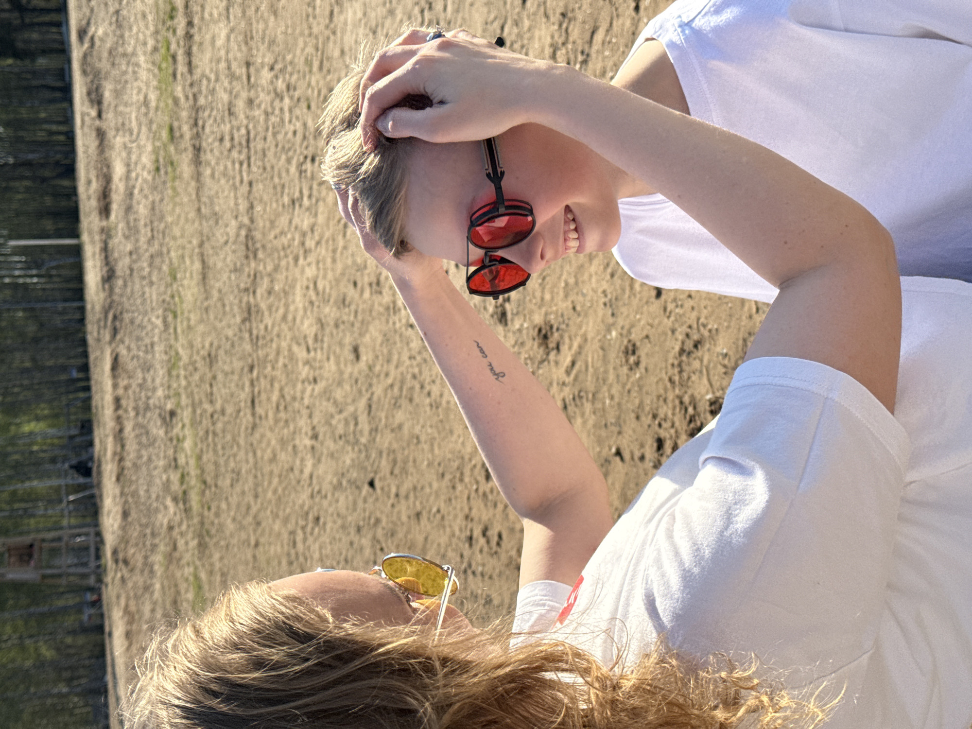 Two individuals are captured in an outdoor setting near the city of Chaikovsky, Russia, on what appears to be a sandy terrain. Both are wearing white t-shirts. The individual closer to the viewer has curly, blonde hair and is wearing yellow sunglasses. The other individual, positioned slightly further away, has short, blonde hair and is wearing red sunglasses, with their hands resting on their head. The background features a fenced area with trees in the distance. The image is rotated counterclockwise.