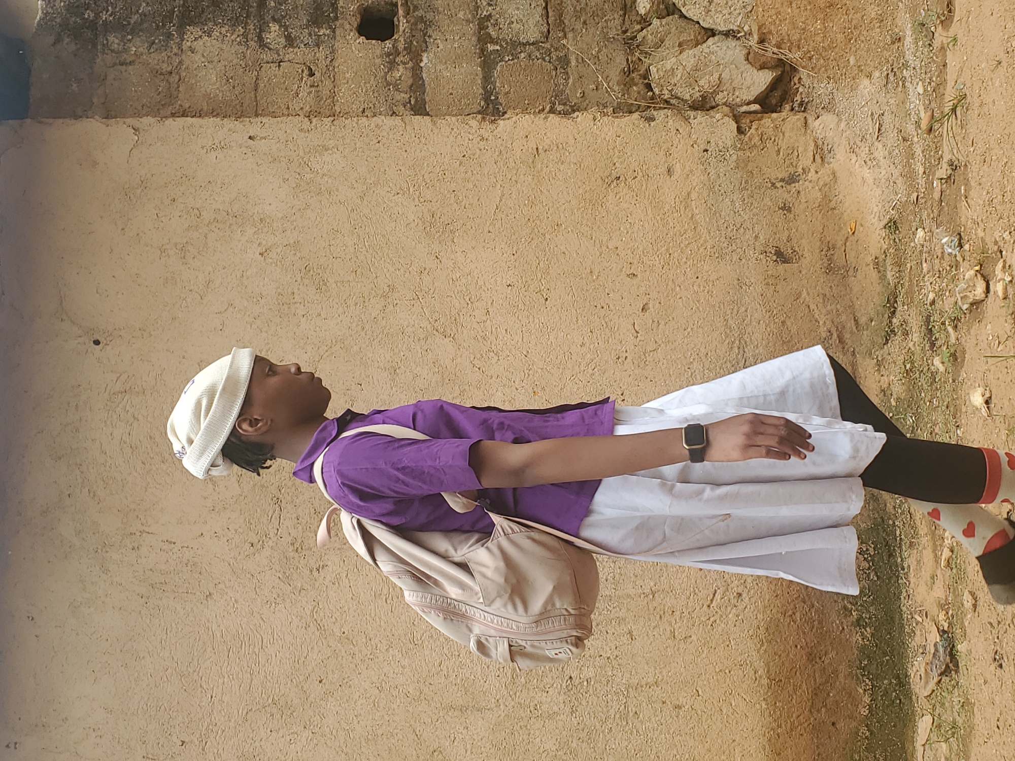 A young person walks past a textured, mud-plastered wall with visible brickwork on the right, suggesting an outdoor urban setting in Jos, Nigeria. The individual is wearing a white knit cap, a purple collared shirt, a white pleated skirt, black leggings, and white socks with red hearts. A light-colored backpack is slung over their left shoulder. They are looking down and to the right, with their right arm bent and hand visible. The ground beneath them is a mix of dirt, small stones, and sparse vegetation. The overall impression is of a person on the move, perhaps commuting, against a backdrop of weathered urban architecture.