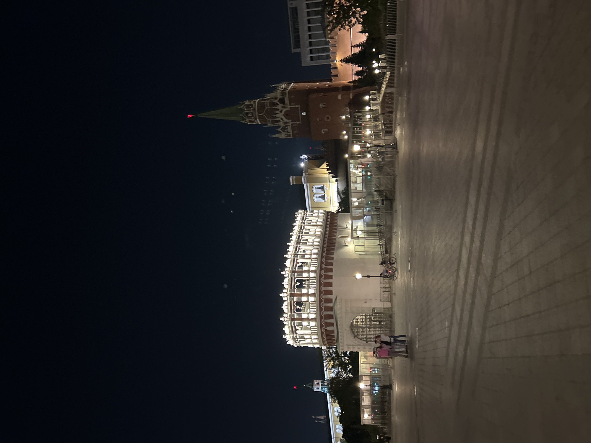 A nighttime exterior shot captures the illuminated facade of a historic building complex. The main structure features a circular tower with arched windows and a red-capped spire, while a long, multi-story building with numerous lit windows runs horizontally to its left. A paved plaza in the foreground is sparsely populated with a few individuals and a bicycle. The scene is illuminated by artificial lights emanating from the buildings and street lamps, casting long shadows across the plaza. The sky is dark and clear.