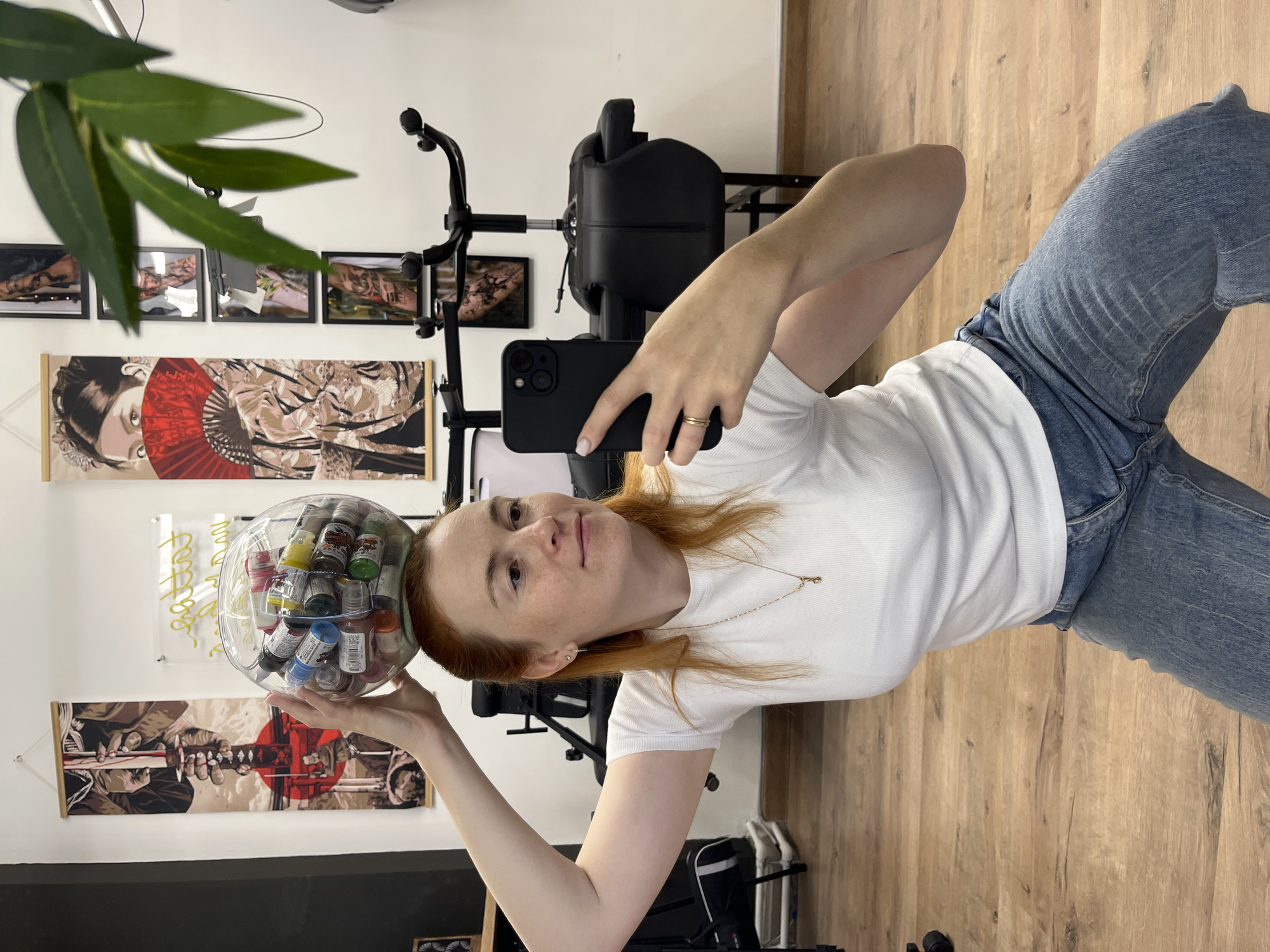 A woman with red hair is taking a selfie in a tattoo studio. She is holding a clear glass bowl filled with small bottles of colorful tattoo ink on top of her head. The woman is wearing a white t-shirt and blue jeans and is looking at her phone. In the background, there is a tattoo chair, a plant, and several pieces of artwork on the wall. The floor is made of wood. The overall impression is that of a lighthearted moment captured within a professional setting.