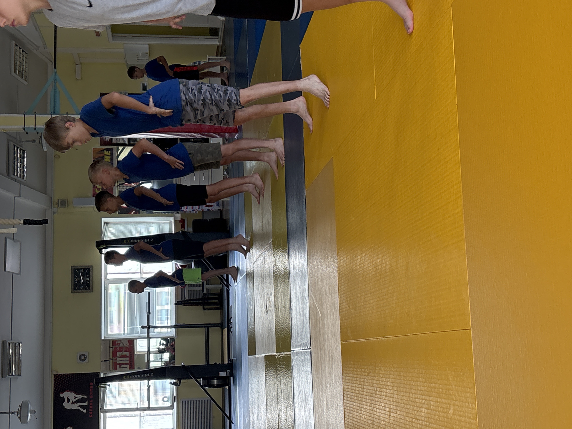 In a gym in Chaikovsky, Russia, a group of young boys stand in a line on a yellow mat, facing away from the camera. They are dressed in athletic wear, with some wearing blue t-shirts and shorts or camouflage-patterned shorts. One boy in the foreground wears a grey t-shirt and black shorts. The gym features various pieces of exercise equipment, including rowing machines and weight racks, along with posters on the wall and a clock. The lighting suggests an indoor setting, possibly during the day.
