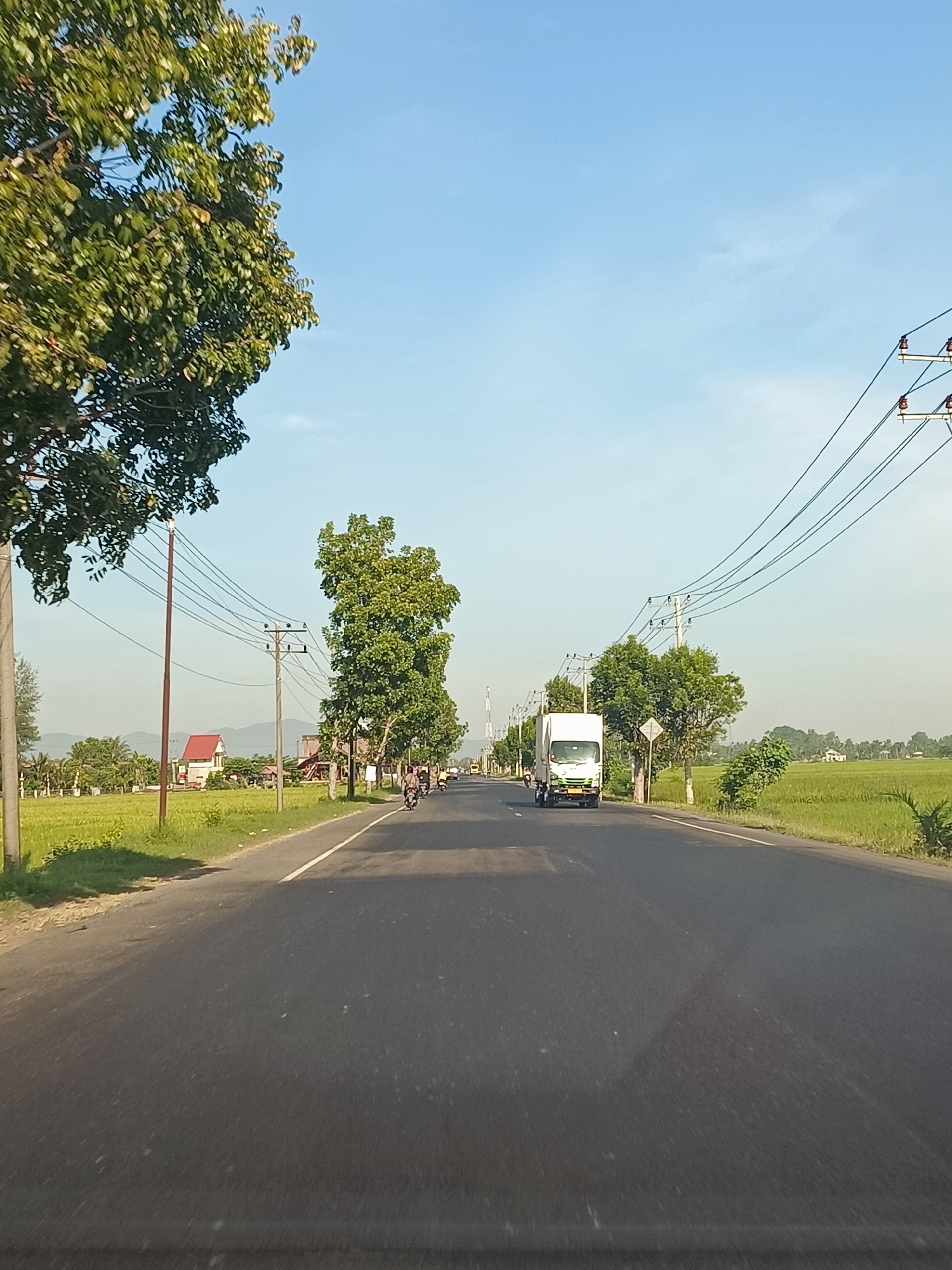 The image depicts a road scene on a clear day, likely in Bili, Indonesia, given the context. The road is paved and appears to be a two-lane street with a white line marking the center. On either side of the road are lush green rice fields, indicative of an agricultural area.
In the foreground, the road surface is visible, with some scattered debris. A slight shadow is cast on the road, possibly from the vehicle the photo was taken from.
Moving along the road, we see various elements. Several motorcycles are present, with riders on them, suggesting daily commuting or local travel. A white truck with a green logo is driving on the road, positioned further down. Utility poles with numerous wires run along the side of the road, a common sight in many urban and rural areas.
Trees are scattered throughout the scene, some with dense foliage and others with sparser leaves. A house with a red roof is visible in the distance, nestled amongst the greenery. The sky is a clear, pale blue, with no clouds in sight. The overall impression is that of a serene, rural or semi-rural environment with active local transportation.