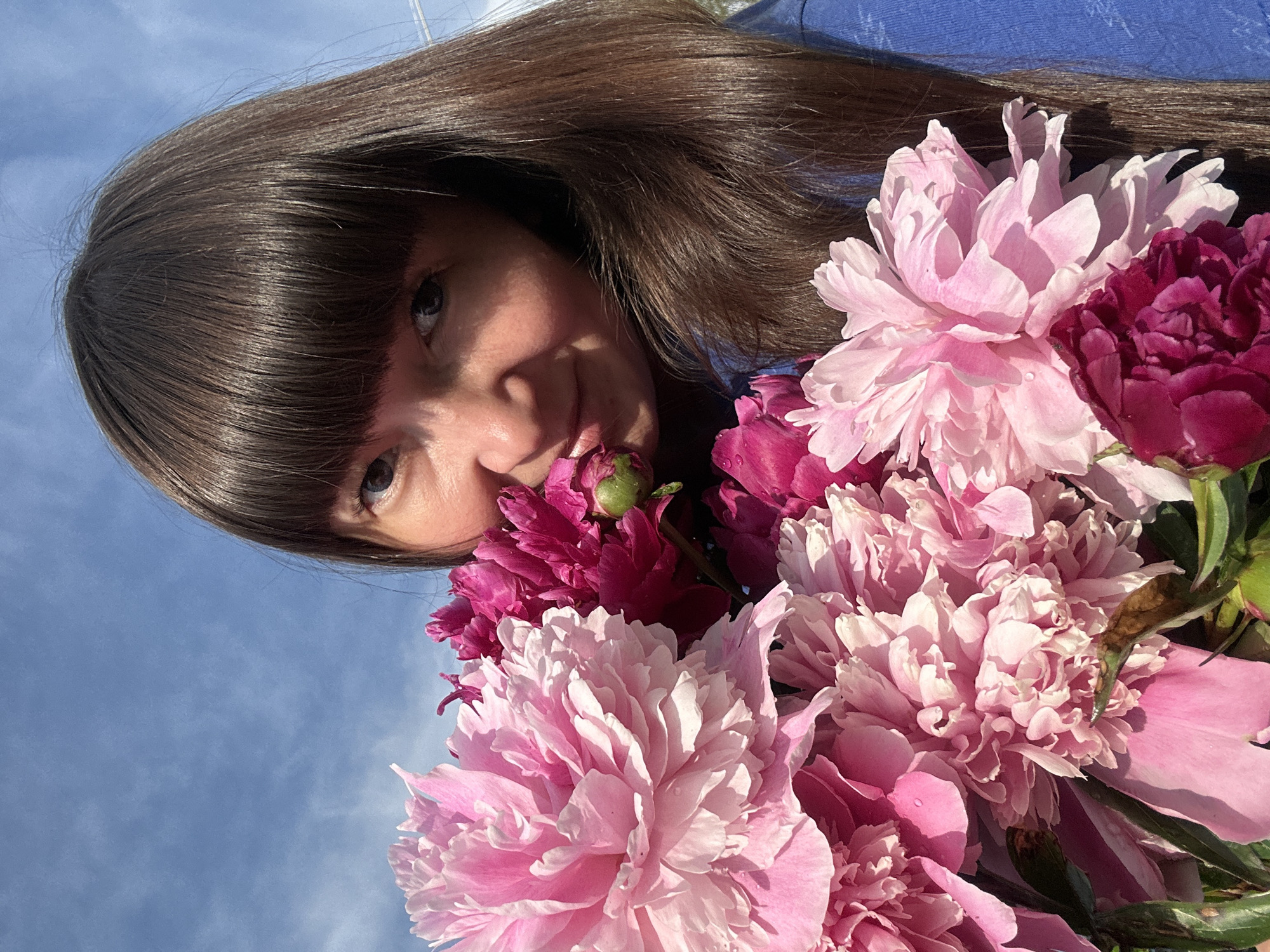 A selfie-style, close-up, eye-level outdoor shot features a woman holding a bouquet of pink and deep magenta peonies. The woman has long, dark brown hair with bangs, and her face is partially obscured by the flowers. She is wearing a blue shirt. The background consists of a bright blue sky with scattered clouds, suggesting it is daytime. The image is taken from a low angle, looking upwards towards the sky. The flowers are the dominant element in the foreground, with petals appearing soft and textured. The scene is likely captured in the vicinity of the village of Sredneye Kechevo, Russia, given the presence of natural flora.