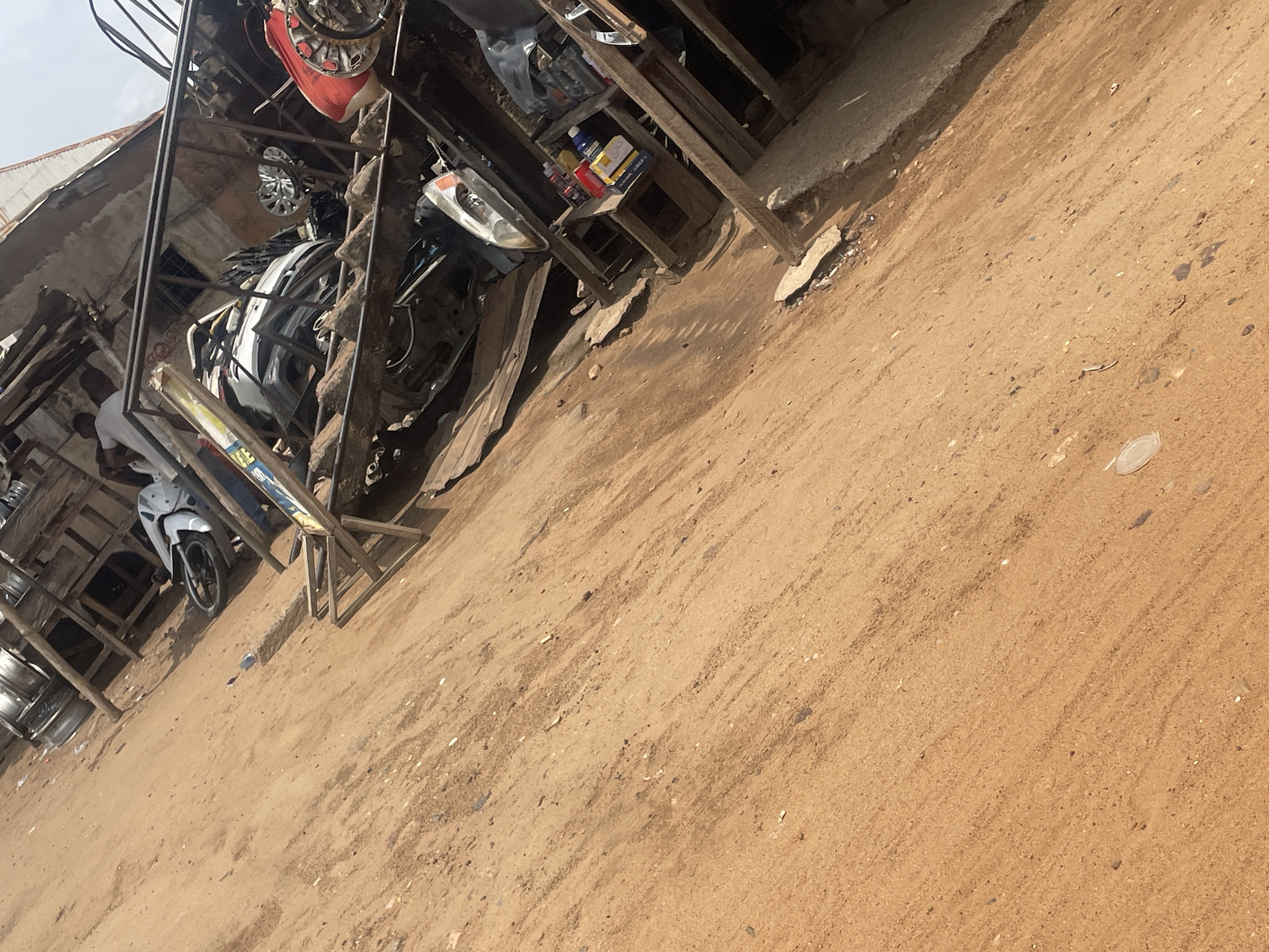 The image depicts an outdoor market scene in Lokoja, Nigeria. The ground is composed of reddish-brown dirt, with scattered stones and debris. In the background, a makeshift structure built from wood and corrugated metal houses various car parts, including a white car chassis, tires, and a red car bumper. A motorcycle is visible to the left, leaning against the structure. Two individuals, both appearing to be men dressed in light-colored clothing, are present near the motorcycle and the car parts. One man is standing, and the other is partially obscured, appearing to be sitting or leaning. The overall scene suggests a repair or salvage yard.