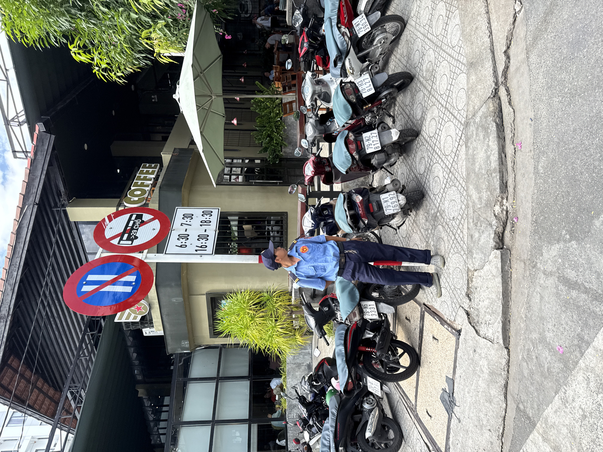 A uniformed male security guard, wearing a blue shirt with a red and gold emblem on the left sleeve, dark pants, and a blue cap, stands facing right on a tiled sidewalk. He has a dark belt with keys attached and is looking off to the right. In front of him, a row of motorcycles is parked, with their seats covered in blue fabric. The motorcycles are of various colors, including black, red, and white. Behind the motorcycles and the guard is the exterior of a building with large glass windows, through which people can be seen seated at tables. A sign above the windows reads "COFFEE" in gold lettering. Two circular red and white traffic signs are visible on a pole to the left of the guard. One sign is a blue circle with a diagonal red bar over two white vertical bars, and the other is a red circle with a diagonal red bar over a black symbol depicting a bus. Below the traffic signs is a white sign with black text indicating parking hours. To the right of the building, a large, light-colored umbrella shades an outdoor seating area. Green foliage is visible above and to the right of the building. The scene appears to be a public space in Nha Trang, Vietnam, likely outside a cafe or restaurant.