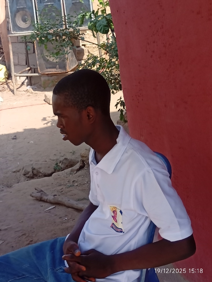 A young man sits outdoors in blue jeans and a white collared shirt. He's positioned to the left, looking away from the camera. To the right is a red wall, while the background features dry ground and some foliage. A large set of speakers is visible in the upper left. The date '19/12/2025 15:18' is stamped in the bottom right corner.