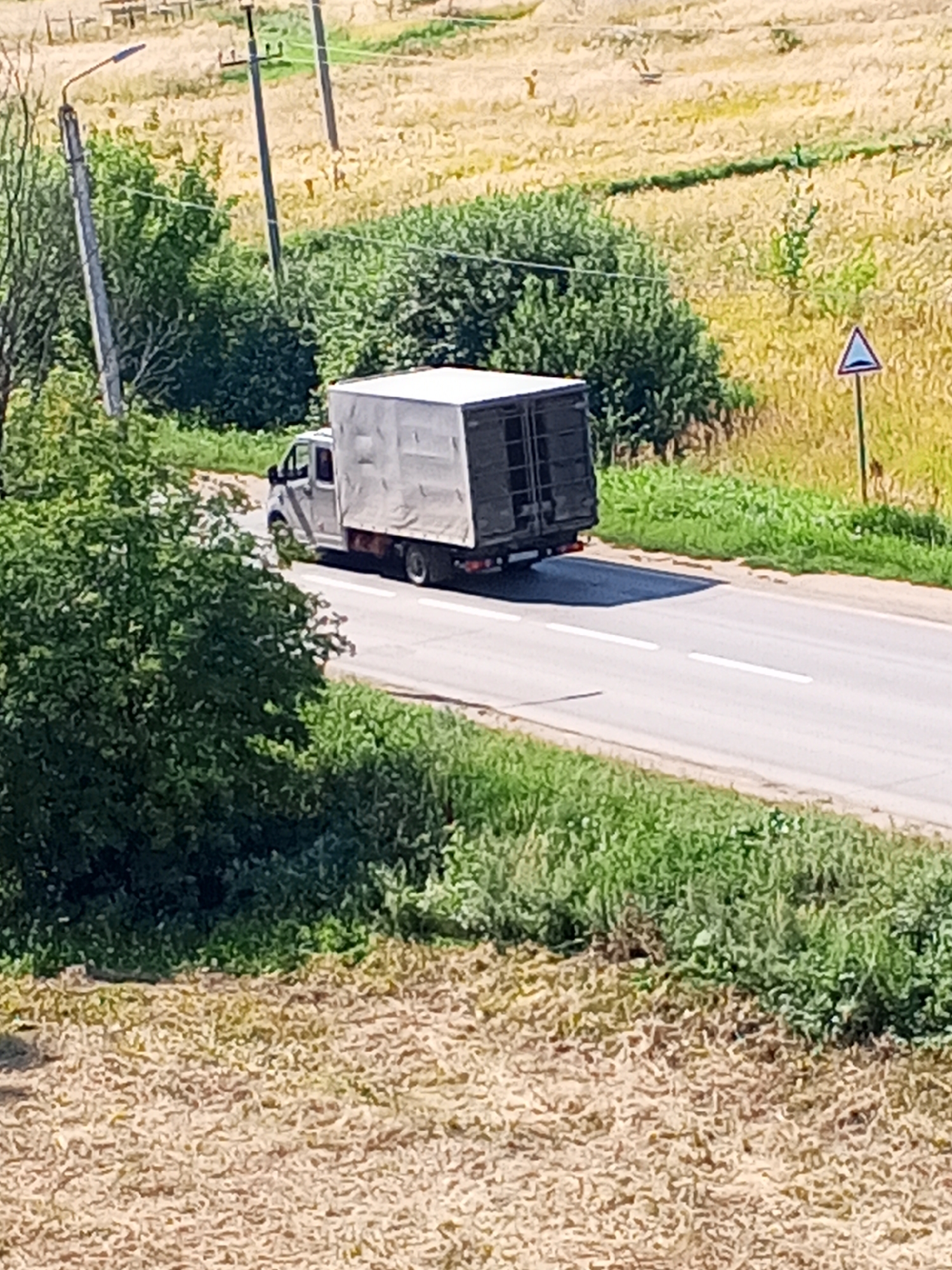 An overcast day in Chistopol, Russia, as a white box truck with a grey tarp covering its cargo drives down a paved road. The truck is on the right side of the road, heading away from the camera. The road is lined with green grass and trees on both sides. In the background, a field of dry, yellow grass stretches to the horizon. A utility pole with a street light stands to the left of the road, and a triangular road sign is visible on the right.