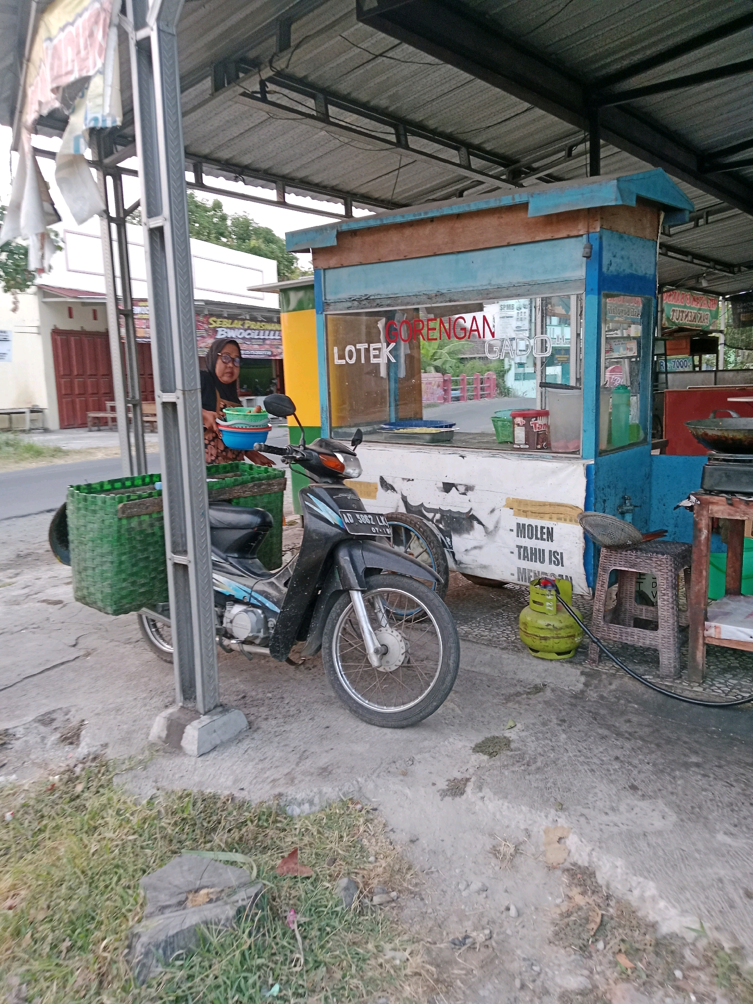This is a full-shot, eye-level, outdoor, vertical, slightly grainy, daytime photograph of a food stall in Waru, Indonesia. The stall is covered by a gray metal roof supported by a gray metal post on the left. A woman wearing a black headscarf and glasses stands behind a blue and light brown food stall, holding two plastic bowls filled with food. The stall has a glass window displaying food items and text in Indonesian that reads "GORENGAN GADO". A black motorcycle is parked in front of the stall, equipped with a large green woven basket on the rear. A green gas cylinder is visible next to the stall, connected to a hose. The ground is paved with concrete and dirt. In the background, a yellow building with red doors and windows is visible. The overall scene depicts a street food vendor preparing for business.
