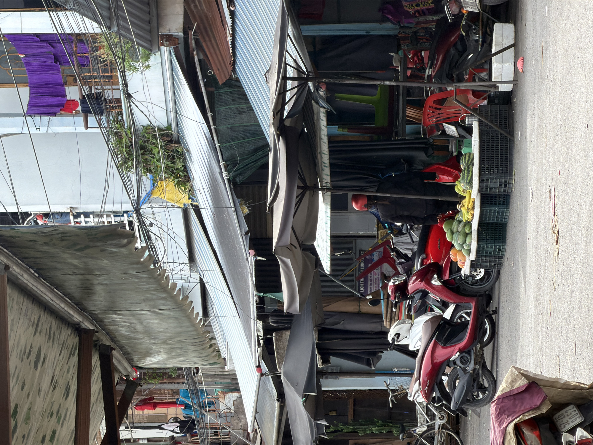 This media file captures a vibrant, sunlit street scene, likely in a market or commercial area of Nha Trang Ward, Vietnam, given its characteristic visual cues. The image is taken from a slightly elevated perspective, looking down a narrow street lined with various stalls and buildings.
**Subjects:** The foreground and middle ground are dominated by several parked motorbikes and scooters of various colors, including red, white, and dark grey. One bicycle is partially visible on the left. On the right, a vendor's stall displays an assortment of fresh fruits like bananas and green mangoes or papayas, arranged in plastic crates. Behind this, colorful plastic chairs (red, green) are stacked, suggesting another stall. A person wearing a dark jacket and a red helmet is visible near the fruit stall, possibly tending to it or their motorbike. The background shows multiple buildings with corrugated metal roofs and awnings. Overhead, a dense network of electrical wires crisscrosses the sky, a common sight in urban Vietnamese landscapes. Purple fabric, possibly laundry, hangs to dry from an upper-story balcony on the left.
**Activity or event taking place:** The scene depicts typical daily commerce and urban life. People are presumably engaged in selling and buying goods, and motorbikes are parked along the street, indicative of transport and daily errands.
**Notable details:** The time of day is clearly daylight, with bright sun casting some shadows, suggesting a clear and possibly warm day. The architecture, the prevalence of motorbikes, and the general market atmosphere strongly confirm the Vietnamese setting. The numerous makeshift awnings and stalls indicate a bustling, informal market environment.
**Visible text:** On a red sign in the background, partially obscured, the words "LICH THI" and "EURO 24" are discernible, likely an advertisement or schedule related to the Euro 2024 football tournament. A motorbike license plate is partially visible with numbers such as "52-Z5" and "603".