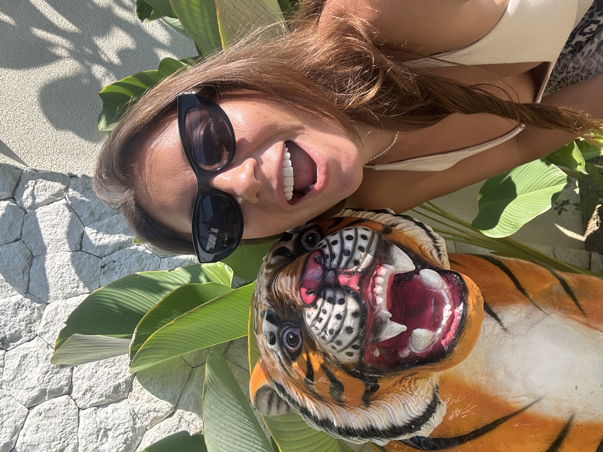 A medium shot shows a woman and a tiger statue outdoors. The woman is lying on her side and looking at the camera. She has long, brown hair, sunglasses, and is wearing a white top. Her mouth is open in a smile or laugh. Next to her is a painted statue of a tiger. The tiger is lying on its side with its mouth open, revealing its teeth and tongue. The tiger's fur is painted orange and black. They are on top of large, green leaves. In the background is a stone wall. This photo was taken in Denpasar, Indonesia.