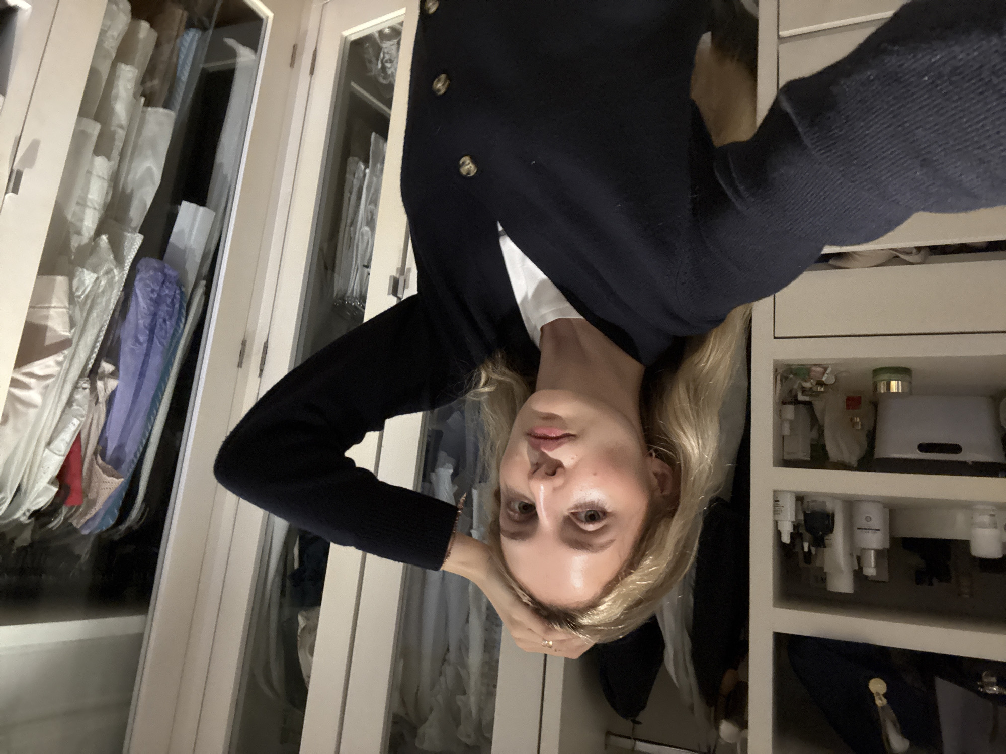 A blonde woman stands in a closet, taking a selfie. She is wearing a navy blue cardigan over a white t-shirt. Her right hand is raised to her head, touching her hair. She has light-colored eyes and fair skin. The closet behind her is filled with clothes on hangers, some of which are visible through glass doors. There are also shelves filled with skincare products and baskets. The setting appears to be a domestic environment. The image does not provide any information or clues about the location being Mexico City, Mexico.