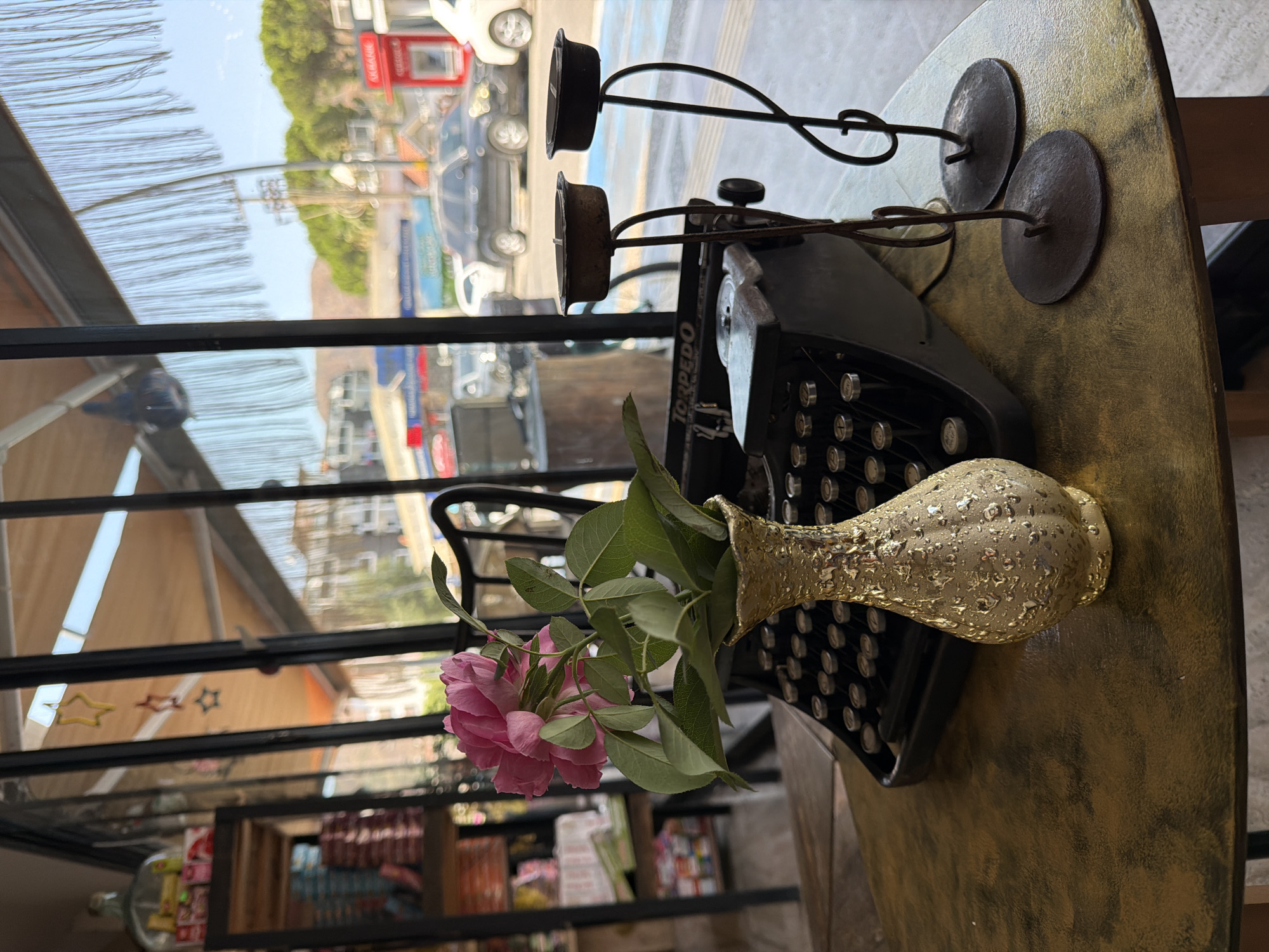 In Marmaris, Turkey, this indoor scene features a gold, textured vase with pink roses and green leaves. The vase sits on a round, metallic gold table next to a vintage black "Torpedo" typewriter. To the right of the typewriter are two wrought-iron candle holders with burnt-out candles. Behind the table, seen through a large window, is a street scene with cars, buildings, and trees. There are also shelves with items on the left, and string decorations hanging from the ceiling.